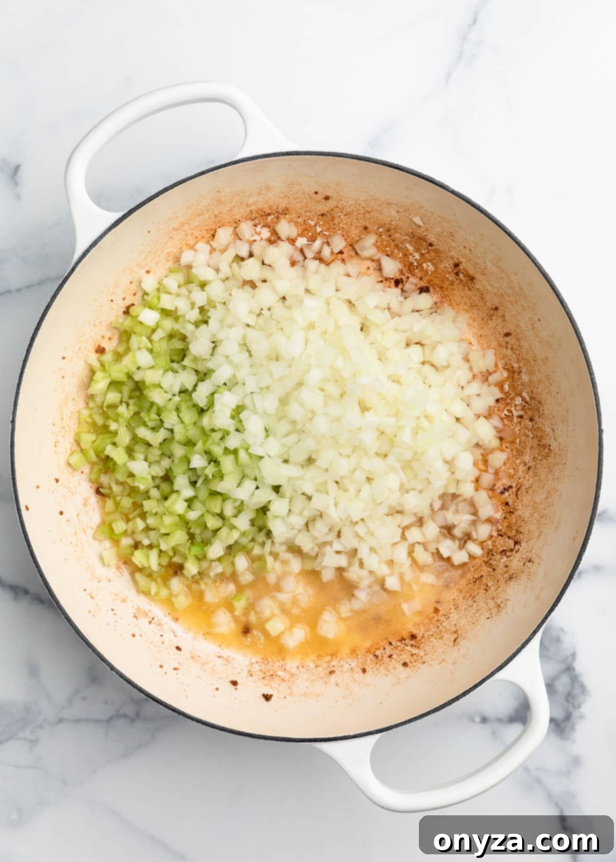 Raw diced celery and onions being added to melted butter and sausage drippings in a white enameled cast iron Dutch oven.