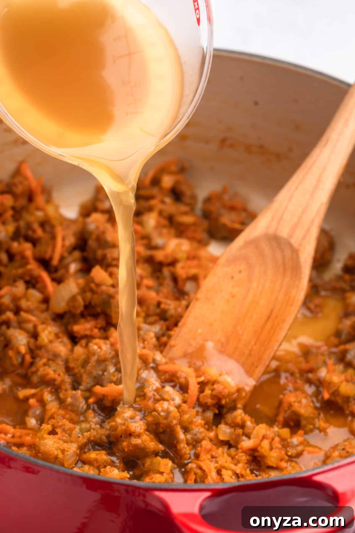 Golden chicken broth being poured gradually into the sautéed sausage and vegetable mixture in a red enameled cast iron Dutch oven, creating the soup's rich base.