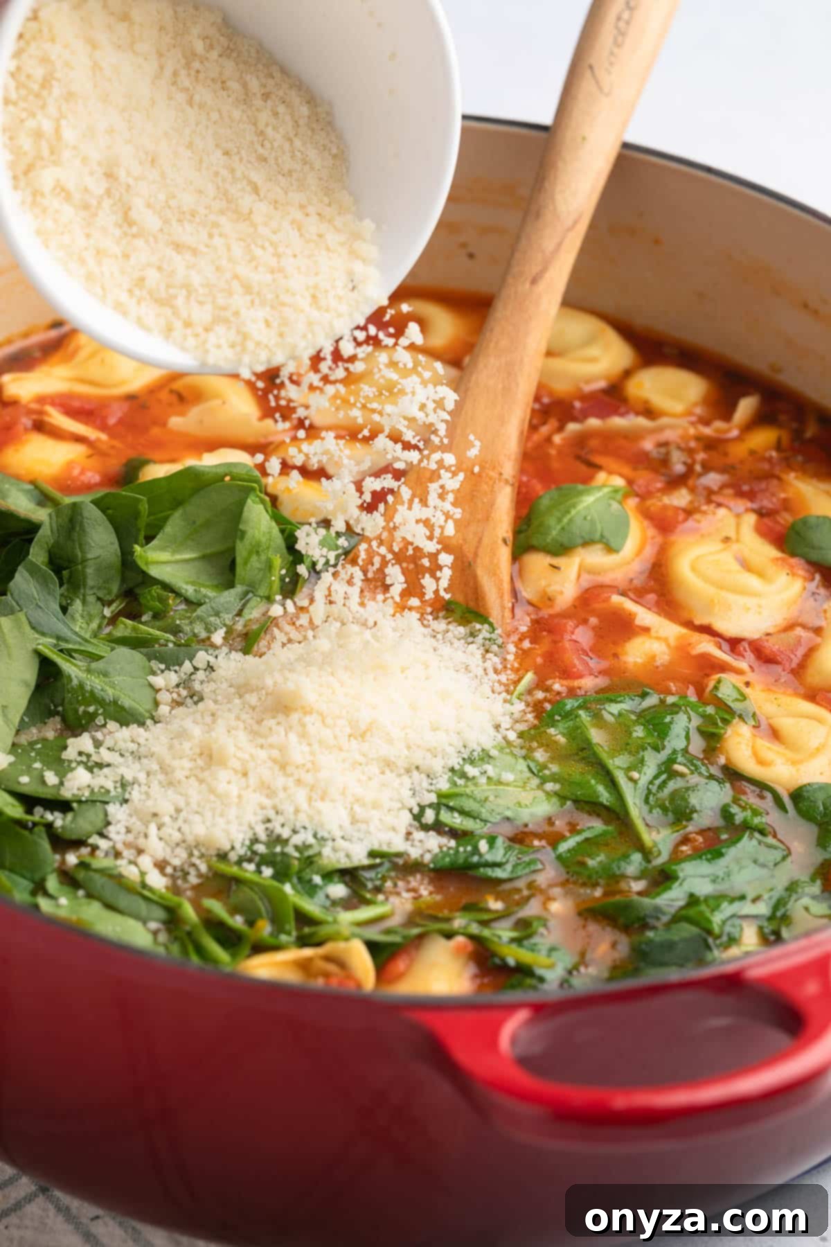 Finely grated Parmigiano Reggiano cheese being generously sprinkled into a pot of Creamy Tortellini Soup, alongside fresh spinach from a white bowl, adding a final layer of rich flavor.
