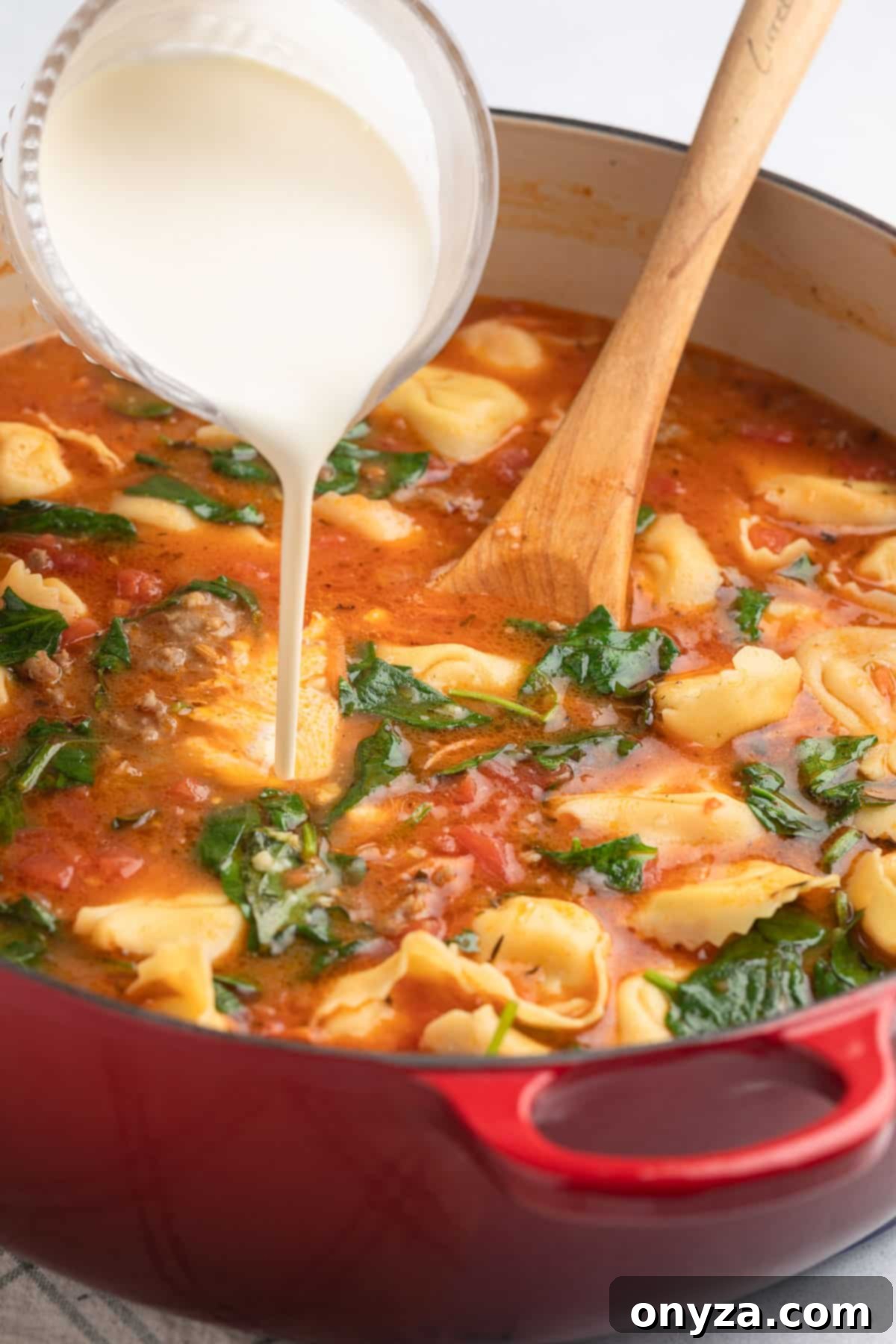 Rich heavy cream being poured from a glass measuring cup into a pot of Creamy Sausage Tortellini Soup with wilted spinach, completing the final creamy texture.