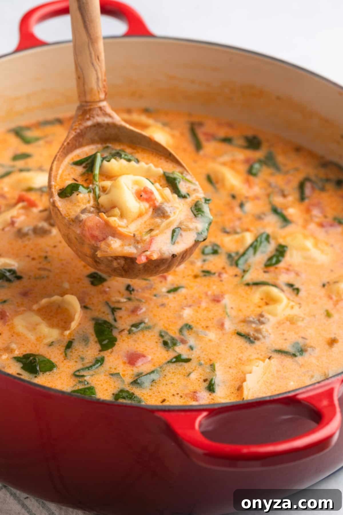 A wooden ladle scooping a generous portion of rich Creamy Sausage Tortellini Soup from a red enameled cast iron Dutch oven, showcasing the hearty ingredients and creamy texture.