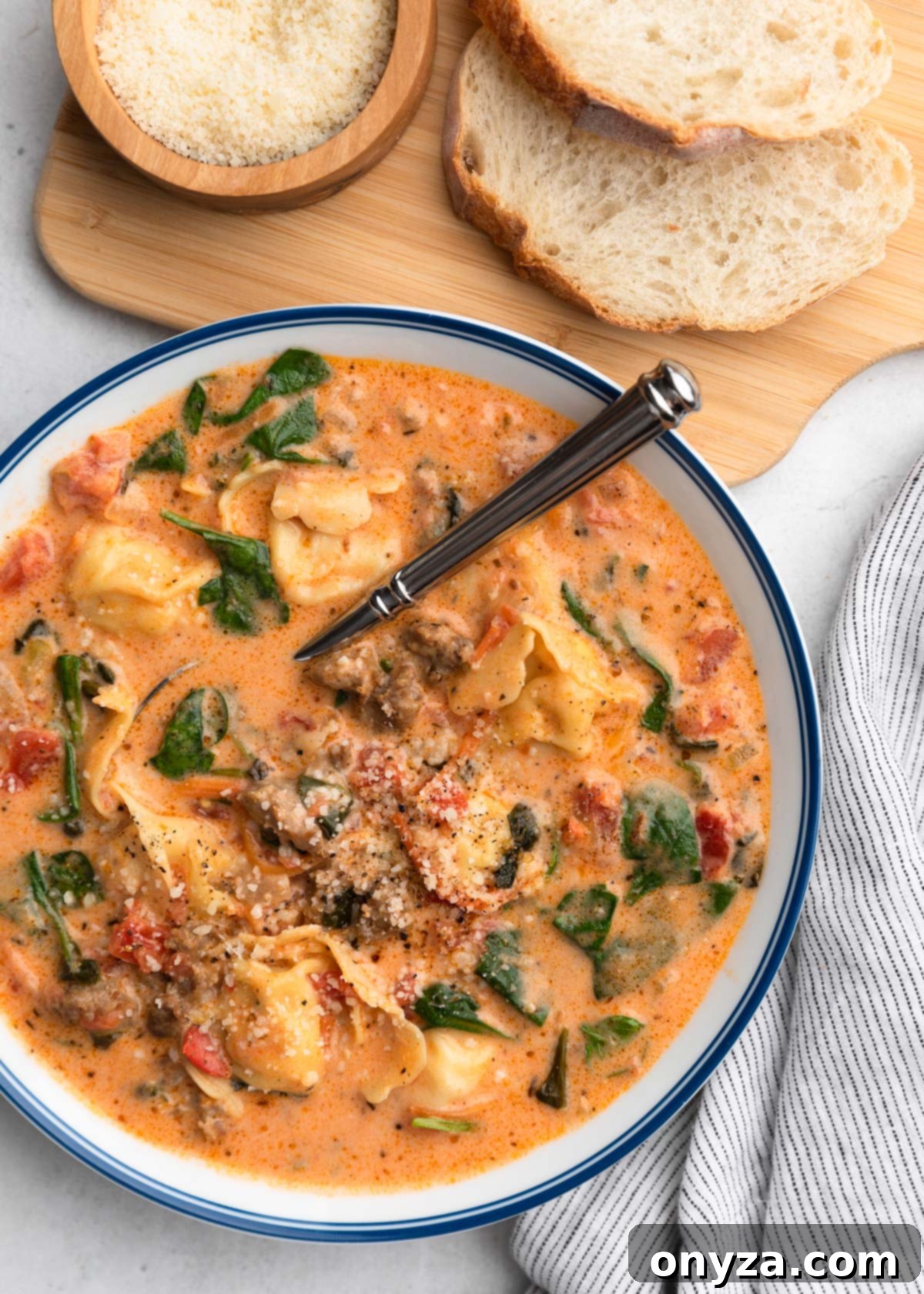 An inviting overhead photo of Creamy Sausage Tortellini Soup in a blue-rimmed white bowl, beautifully presented alongside a wooden serving paddle with slices of crusty Italian bread and a small bowl of grated Parmesan cheese.
