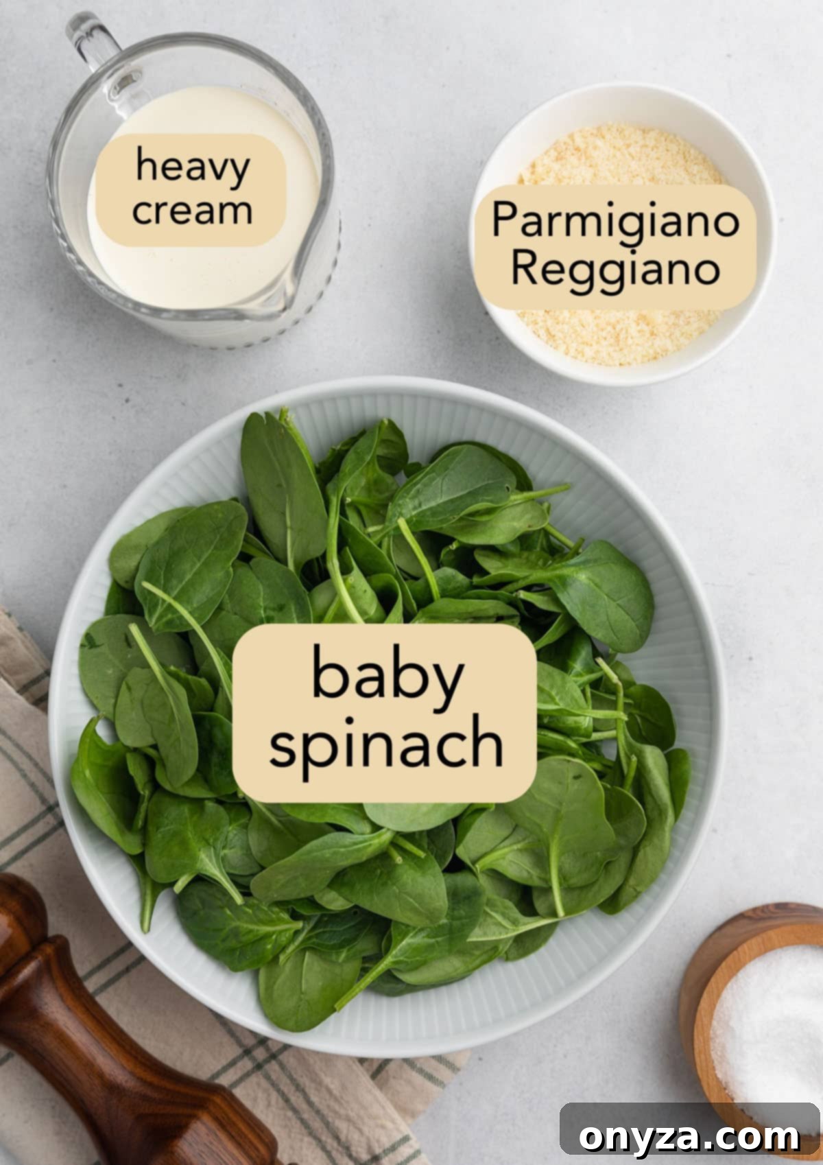 Overhead shot of essential finishing ingredients for Creamy Sausage Tortellini Soup: a bowl of fresh baby spinach, a cruet of rich heavy cream, a bowl of finely grated Parmesan cheese, and a small dish of salt, all on a crisp white background.