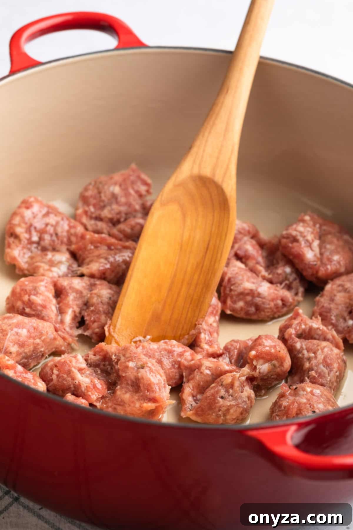 Raw sweet Italian sausage being broken up in a red enameled cast iron Dutch oven with a wooden spoon, ready for browning.