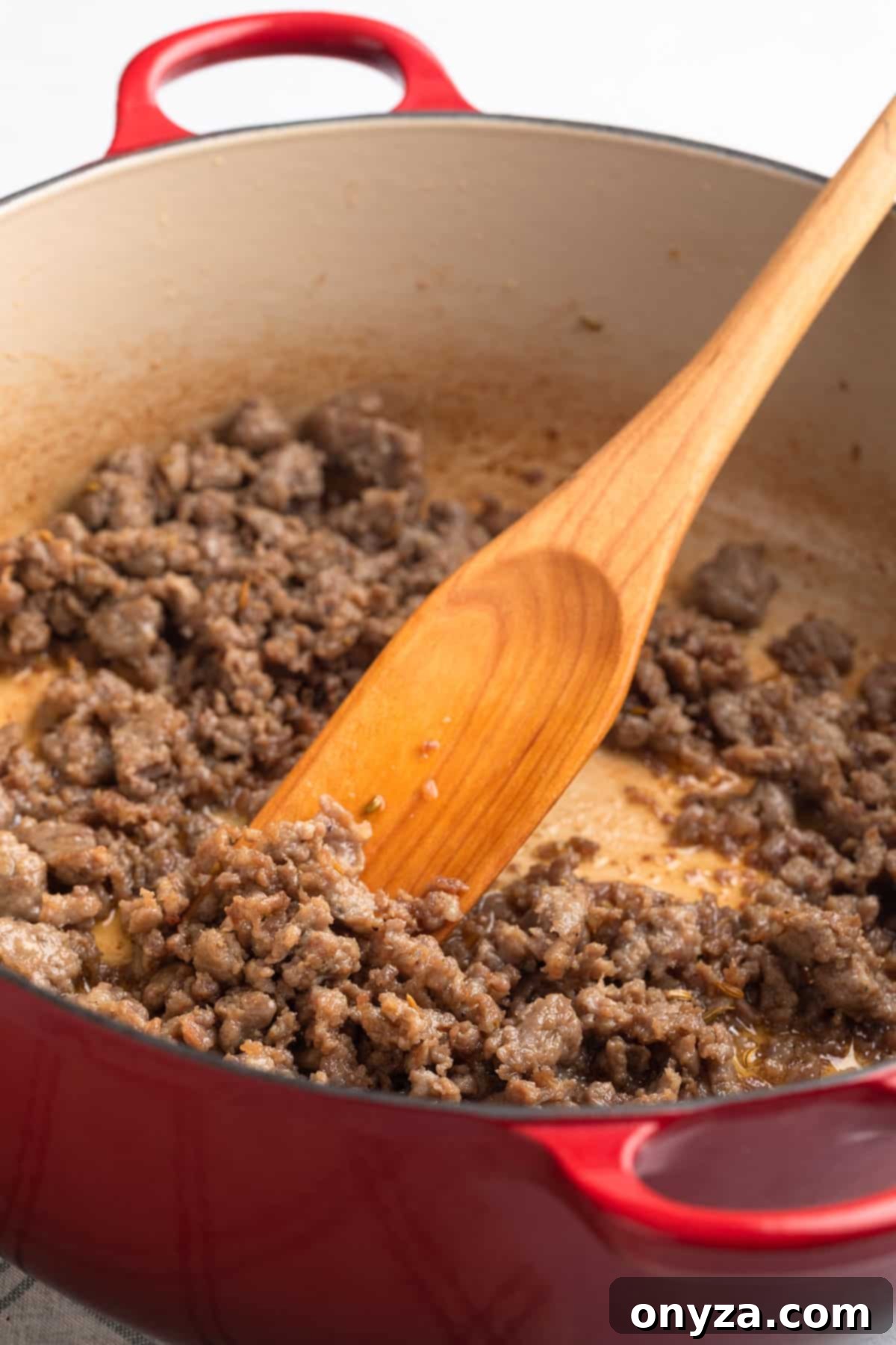 Perfectly browned and crumbled Italian sausage resting in a red enameled cast iron Dutch oven with a wooden spoon, ready to be removed.