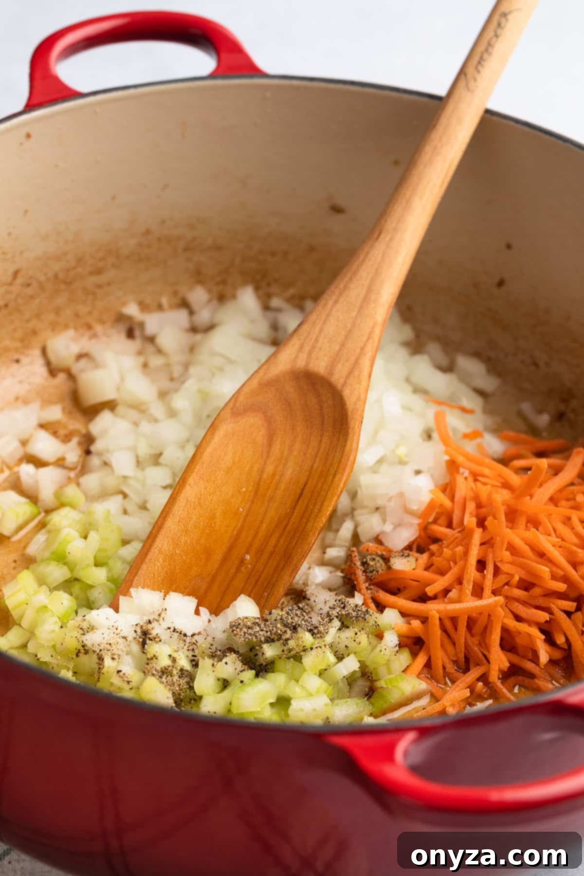 A colorful medley of chopped onions, celery, and carrots in a red enameled cast iron Dutch oven with a wooden spoon, poised for sautéing and building the soup's flavor.