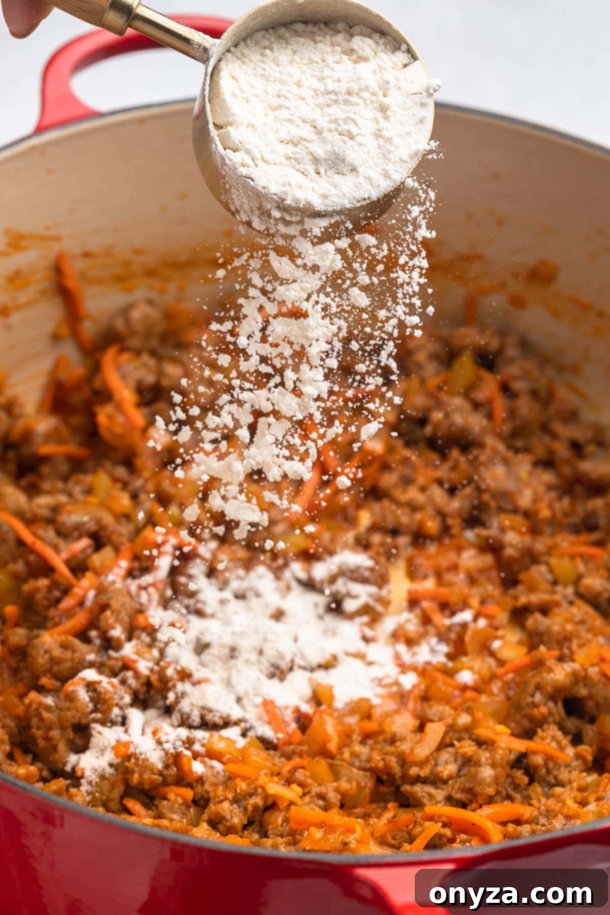 All-purpose flour being sprinkled into browned Italian sausage and sautéed aromatics in a red enameled cast iron Dutch oven, preparing to create a velvety roux.