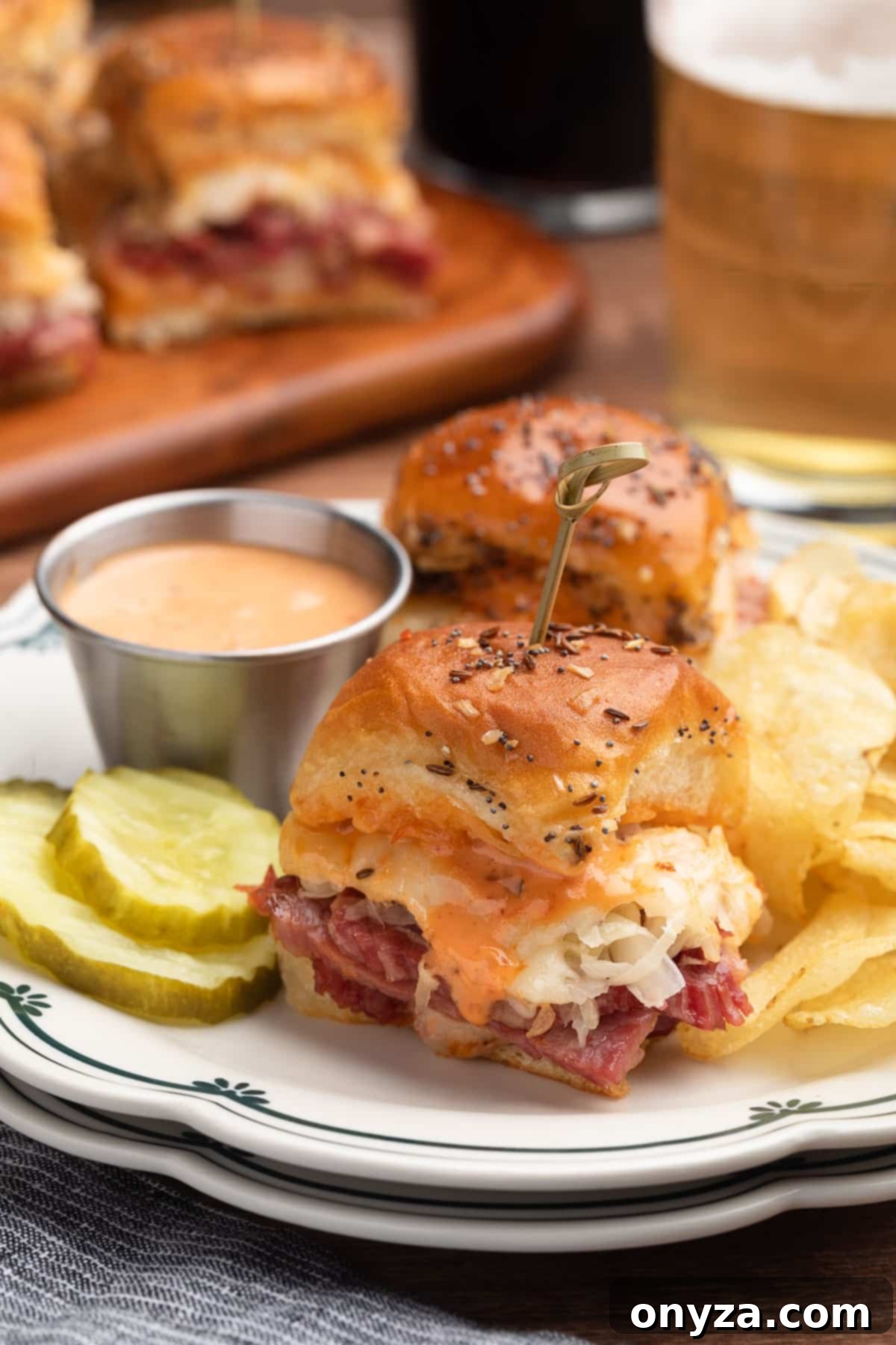 two Reuben sliders on a scalloped white ceramic dish, with potato chips, pickles, and a dipping cup of Russian dressing