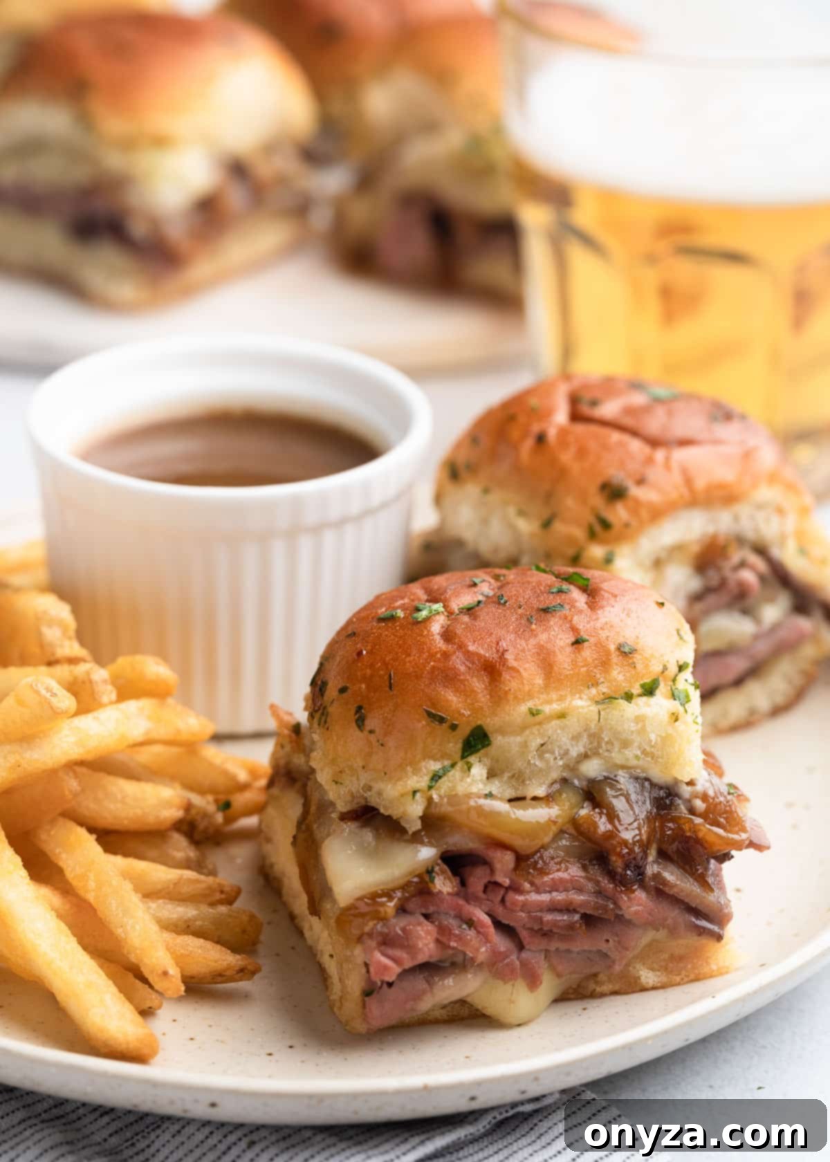 Savory French Dip Sliders 2 french dip sliders on an ivory ceramic plate with french fries and a cup of au jus