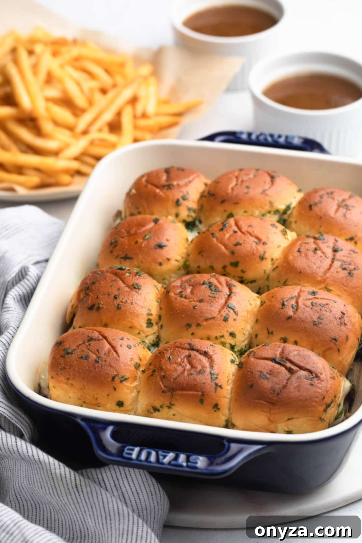 Savory French Dip Sliders 12 baked french dip sliders in a white and blue baking dish with bowls of au jus and a plate of french fries