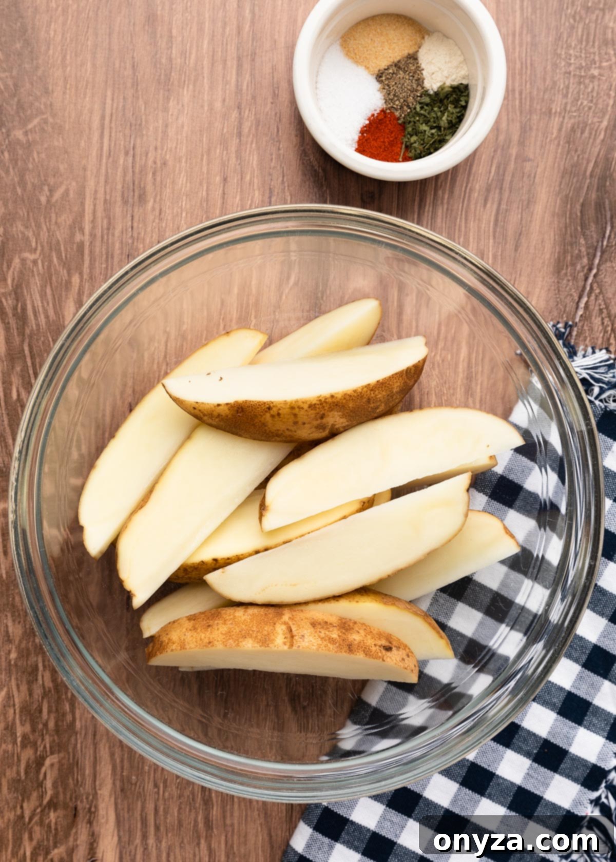 wedged russet potatoes in a glass bowl next to a blue and white check napkin and a smaller bowl of ground spices.