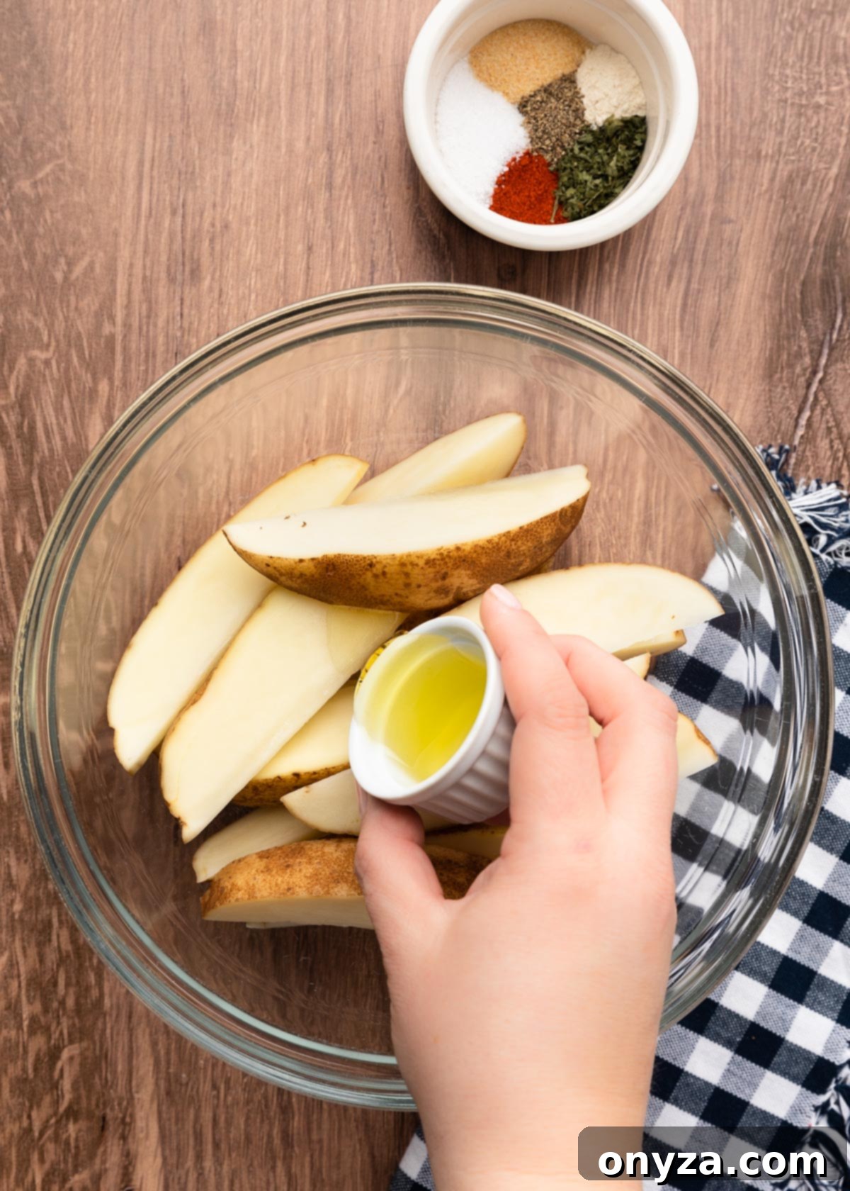 pouring olive oil from a tiny white ramekin into a bowl of wedged russet potatoes