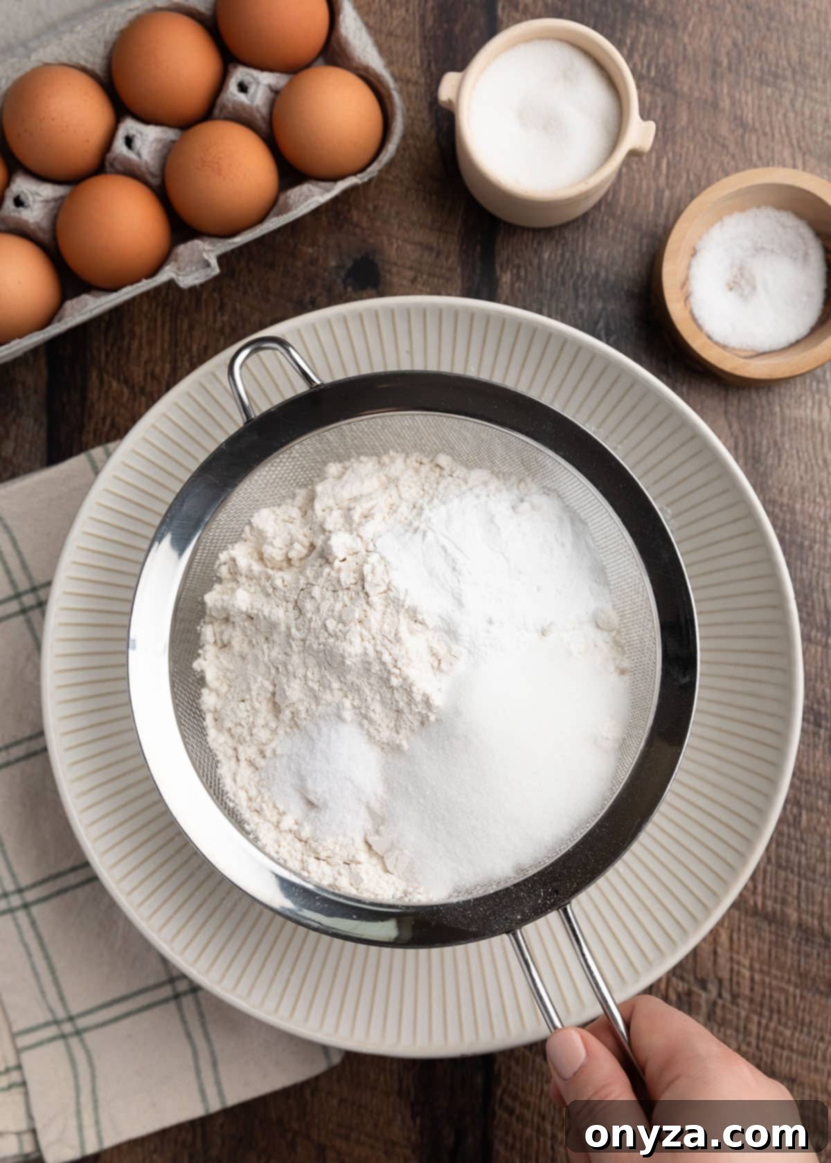 overhead photo of flour, sugar, salt, and baking powder being sifted into a bowl with a fine mesh sieve