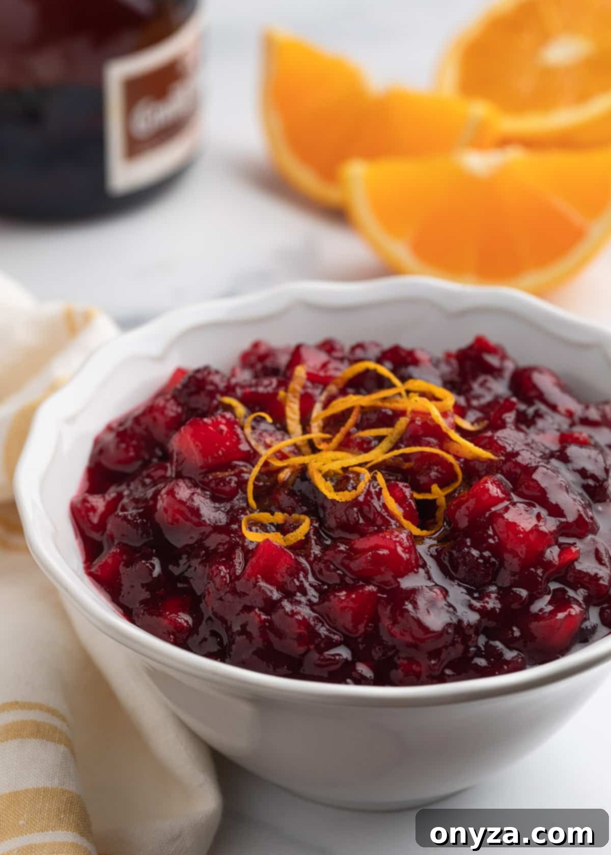 Orchard Citrus Cranberry Sauce 2 A glistening bowl of homemade apple orange cranberry sauce, garnished with fresh orange wedges, sits beside a charming yellow and white striped towel. In the background, a bottle of orange liqueur hints at an optional festive touch.
