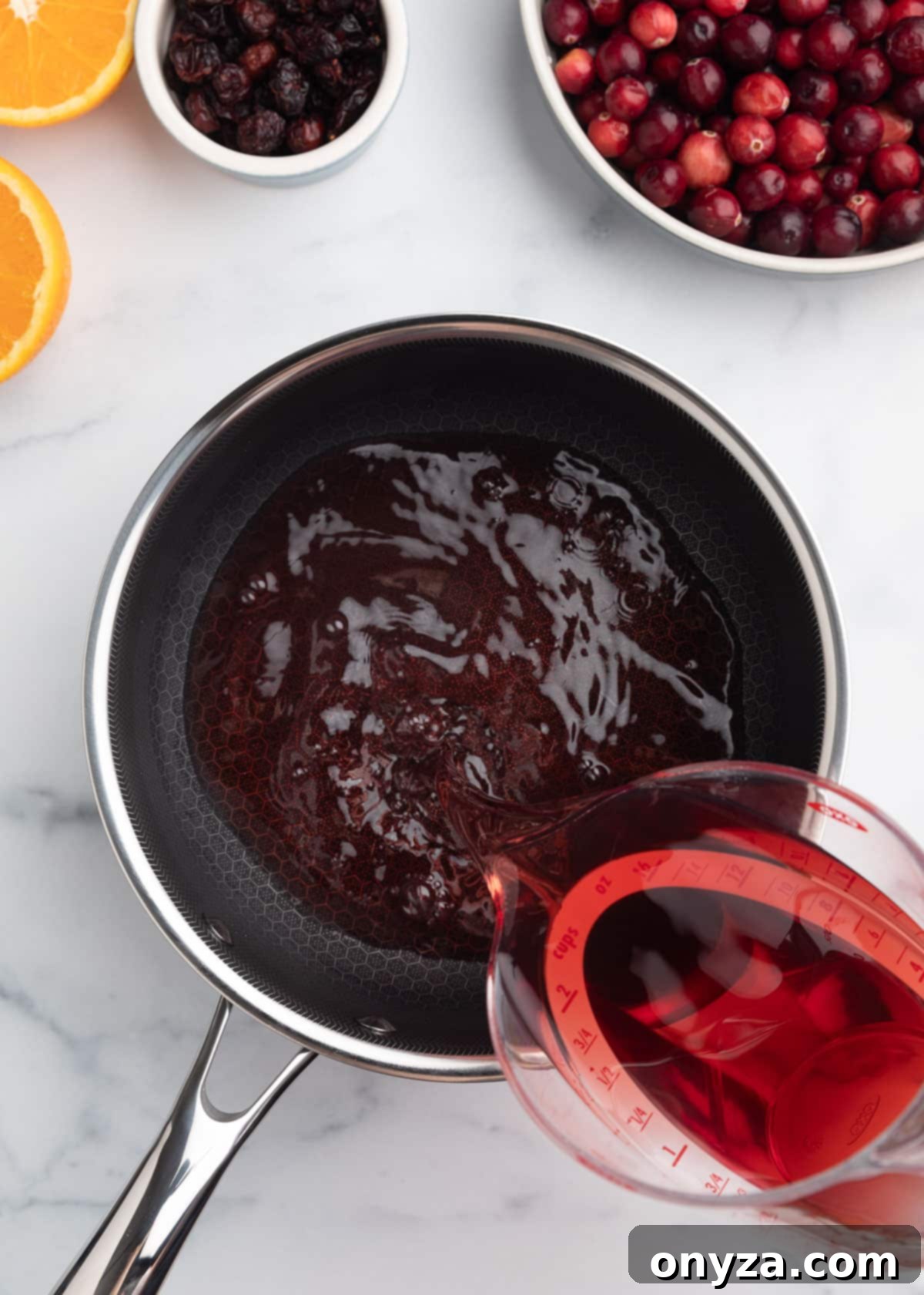 Orchard Citrus Cranberry Sauce 4 An overhead view captures cranberry juice cocktail being carefully poured from a liquid measuring cup into a gleaming saucepan, resting on a elegant white marble board. The setup is ready for the first step of the sauce preparation.
