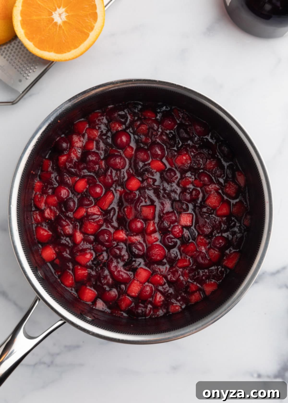 Orchard Citrus Cranberry Sauce 6 An inviting overhead shot reveals the apple cranberry sauce simmering gently in a saucepan on a polished marble board, steam subtly rising from the rich, bubbling mixture. The cranberries have burst, and the apples are tender.