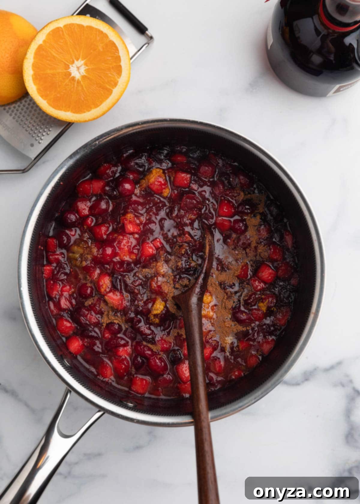 Orchard Citrus Cranberry Sauce 8 A wooden spoon is actively stirring vibrant orange marmalade, fresh orange juice, aromatic zest, and warming ground spices into a rich cranberry sauce within a saucepan. The mixture promises a harmonious blend of sweet, tart, and spiced notes.
