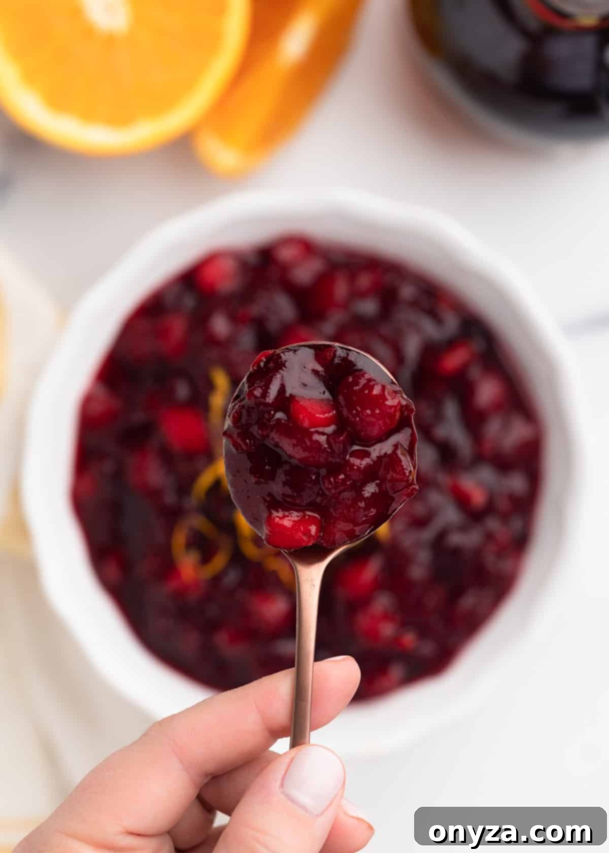 Orchard Citrus Cranberry Sauce 9 An artfully captured overhead view shows a spoonful of glistening apple orange cranberry sauce being held delicately over a pristine white bowl filled with the vibrant sauce. This highlights the sauce's luscious texture and rich color.