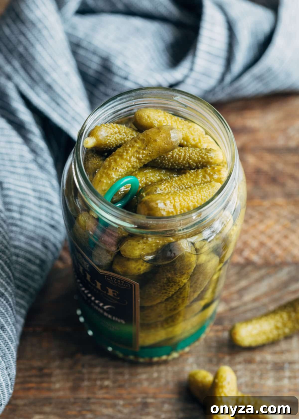 A glass jar filled with green French cornichons, sitting on a rustic wooden board next to a light blue napkin, evoking a sense of gourmet simplicity and traditional culinary charm.
