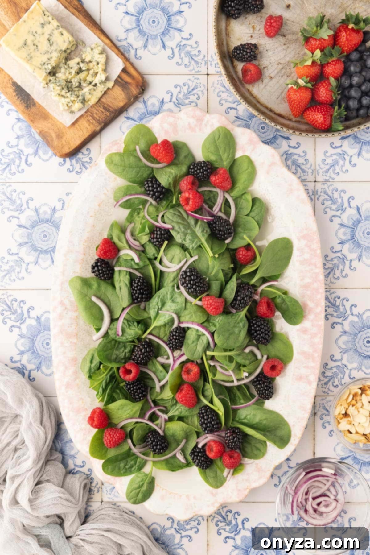 Overhead shot showcasing a vibrant mix of baby spinach, thinly sliced red onions, juicy blackberries, and sweet raspberries arranged on an oval ivory platter against a blue and white tile background.