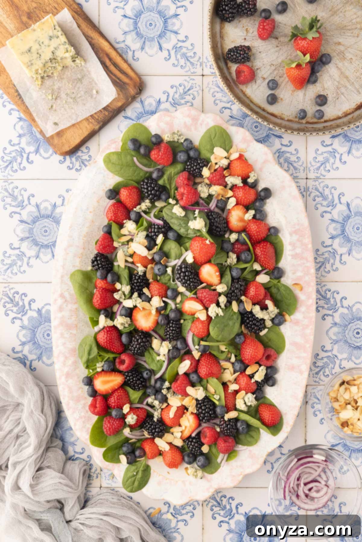 Overhead view of a prepared baby spinach and berry salad, featuring crumbled blue cheese and toasted almonds, awaiting dressing on an ivory oval platter. The backdrop is a stylish blue and white tile surface.