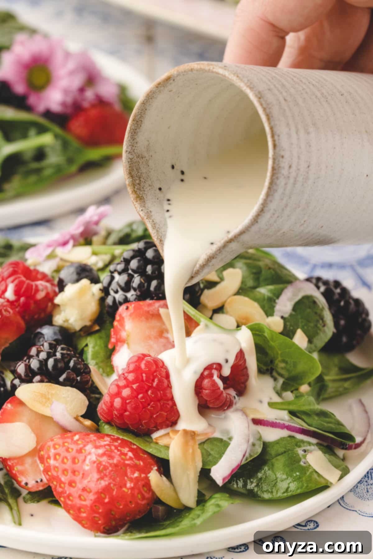 A delicate stream of creamy poppy seed dressing being poured from a pristine white creamer over a freshly assembled spinach and berry salad.
