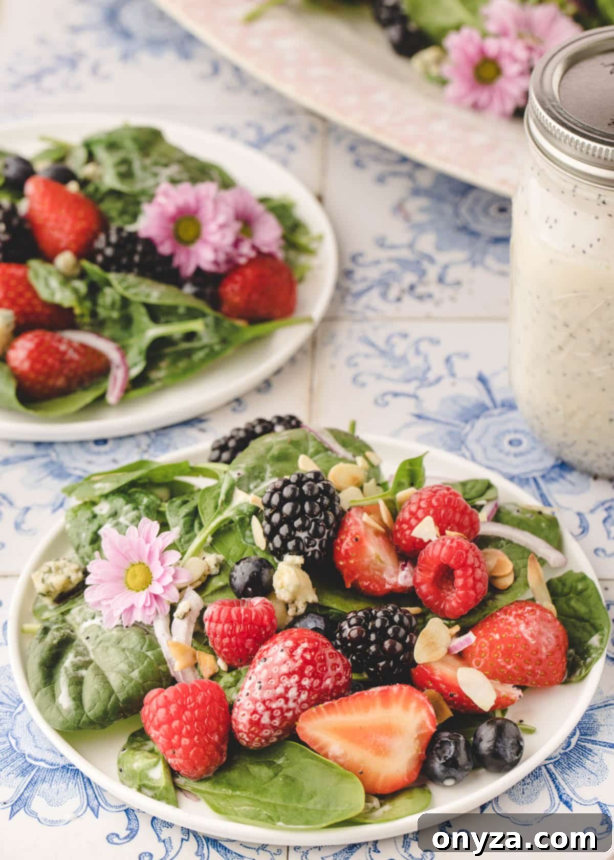 Two elegantly plated portions of spinach berry salad resting on a blue and white tiled surface, with a mason jar of creamy poppy seed dressing nearby.