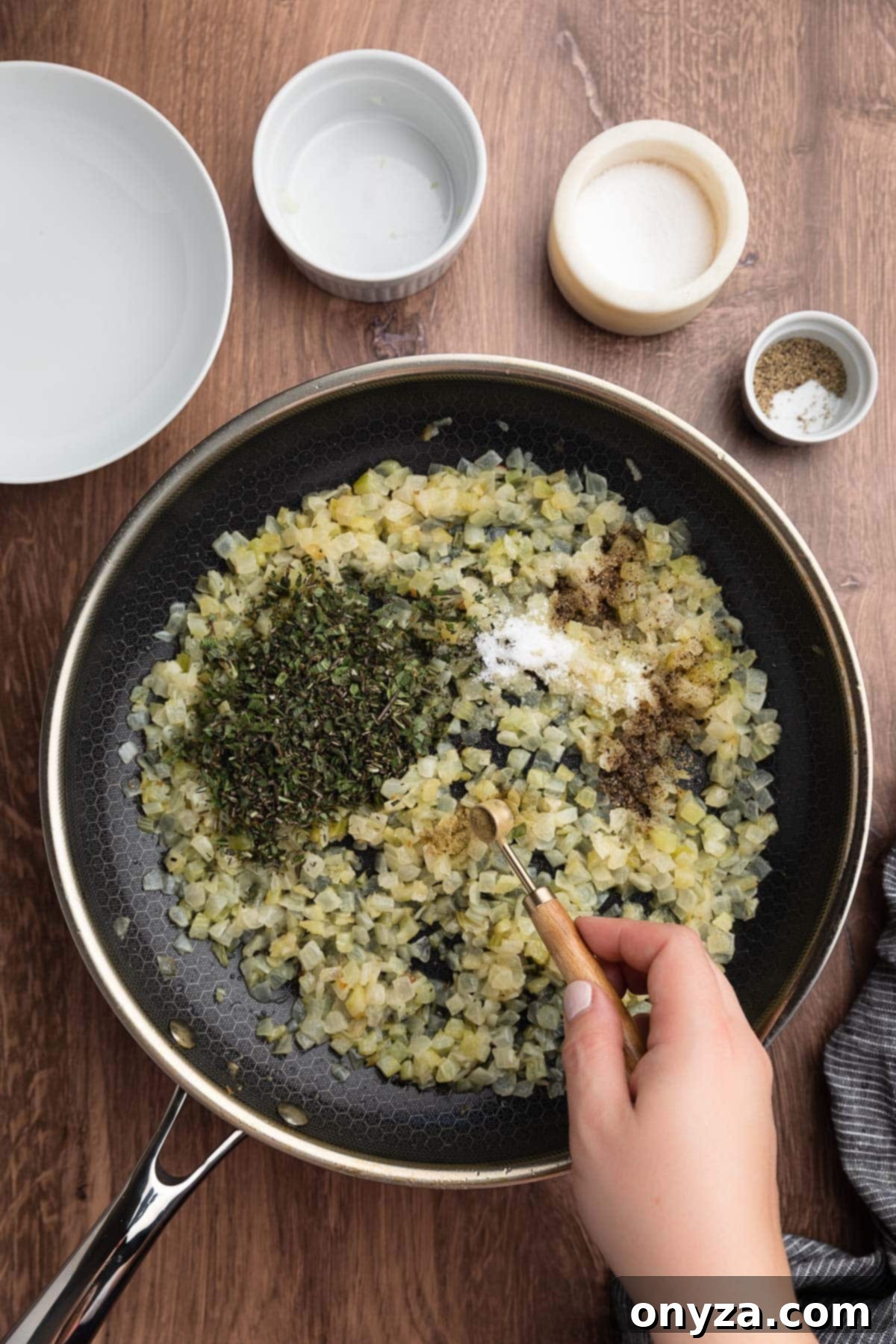 Festive Sausage and Chestnut Stuffing 10 poultry seasoning being added from a dry measuring spoon to a skillet of cooked onions and celery with fresh herbs, salt and pepper.