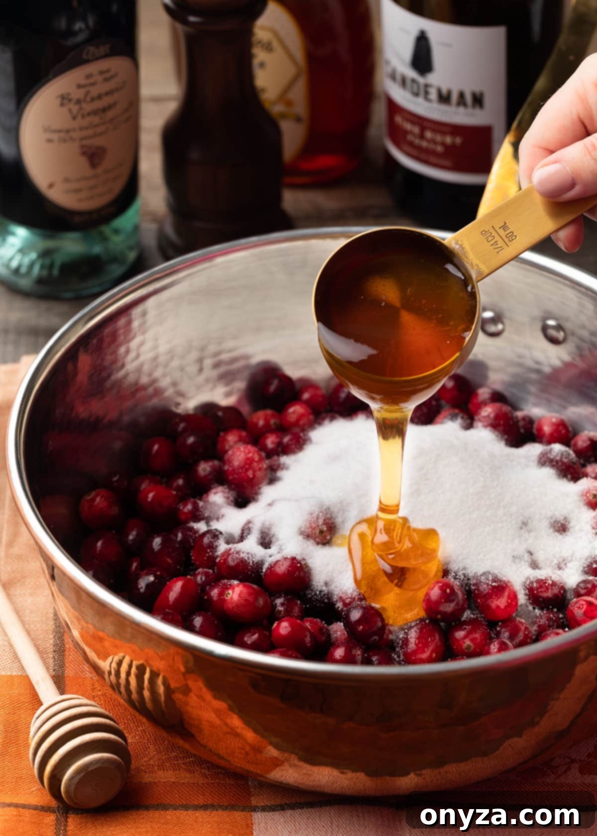 pouring honey from a gold-toned measuring cup into a copper saucepan filled with fresh cranberries and granulated sugar