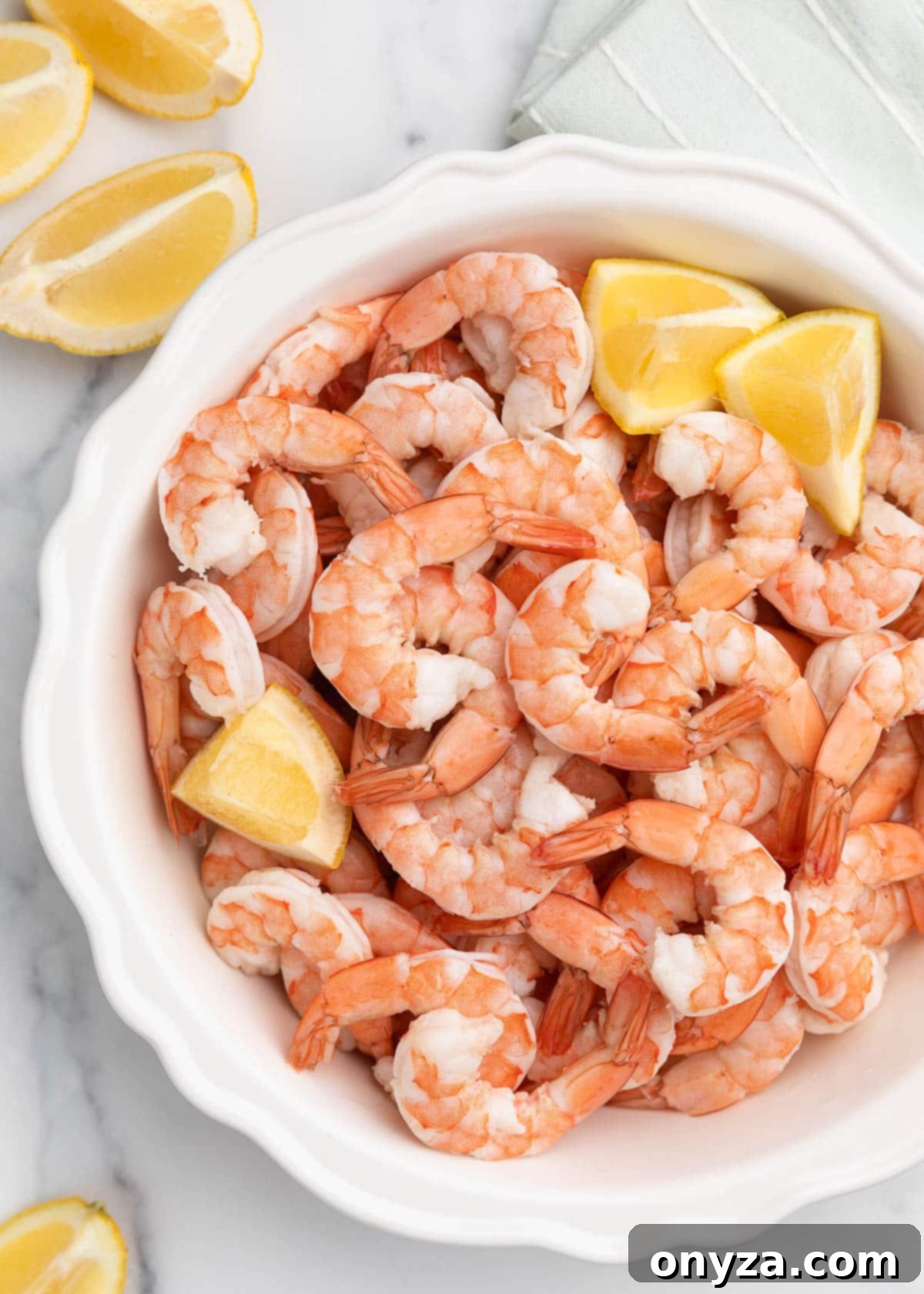 Overhead view of perfectly poached shrimp arranged in a white fluted pie plate, garnished with fresh lemon wedges, all resting on a elegant marble board.