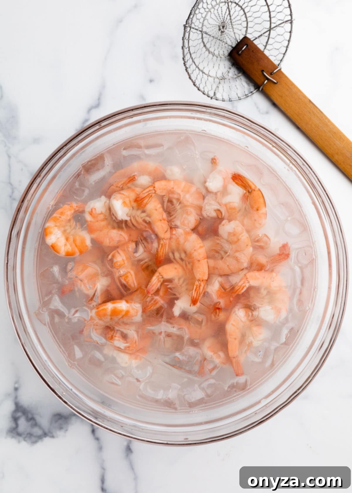 Overhead view of freshly poached shrimp cooling in an ice bath in a large glass bowl, positioned next to a spider strainer on a white marble board.