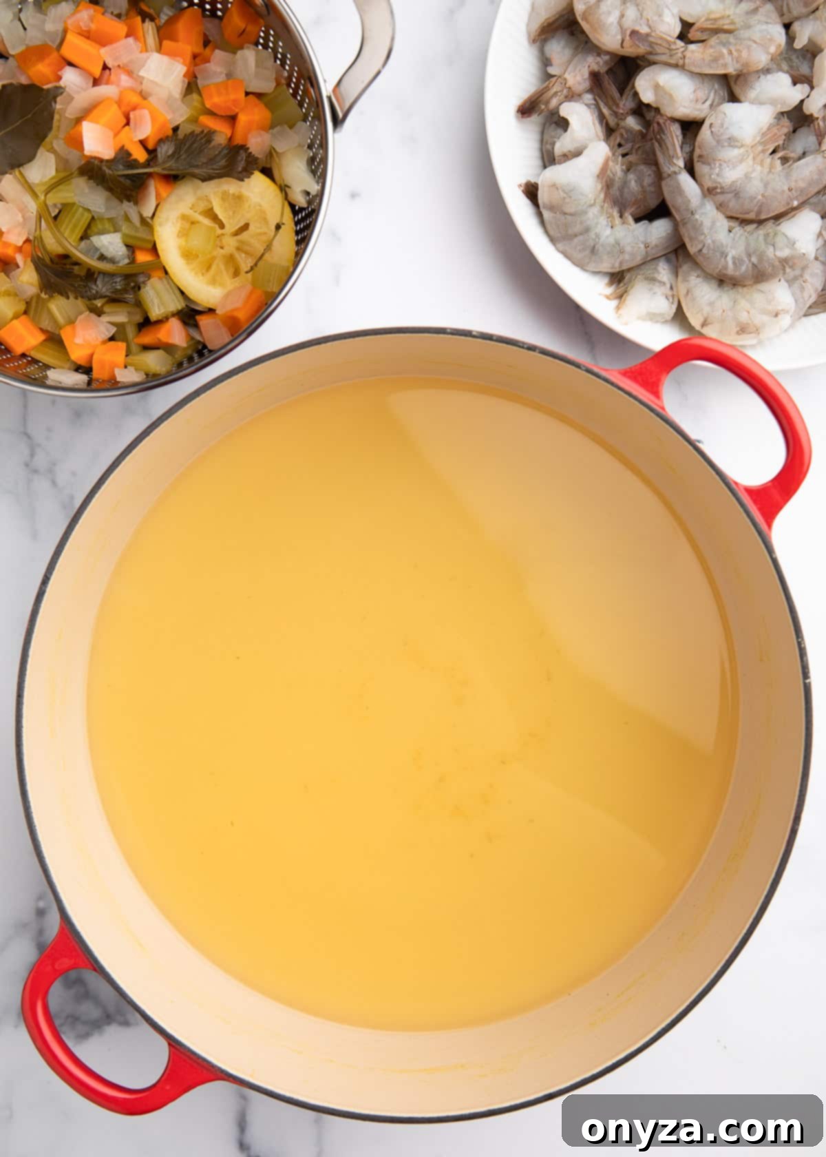 Overhead view of strained court bouillon in a cast iron Dutch oven, positioned next to a colander containing the discarded cooked vegetables and herbs, with a plate of raw shrimp waiting nearby.