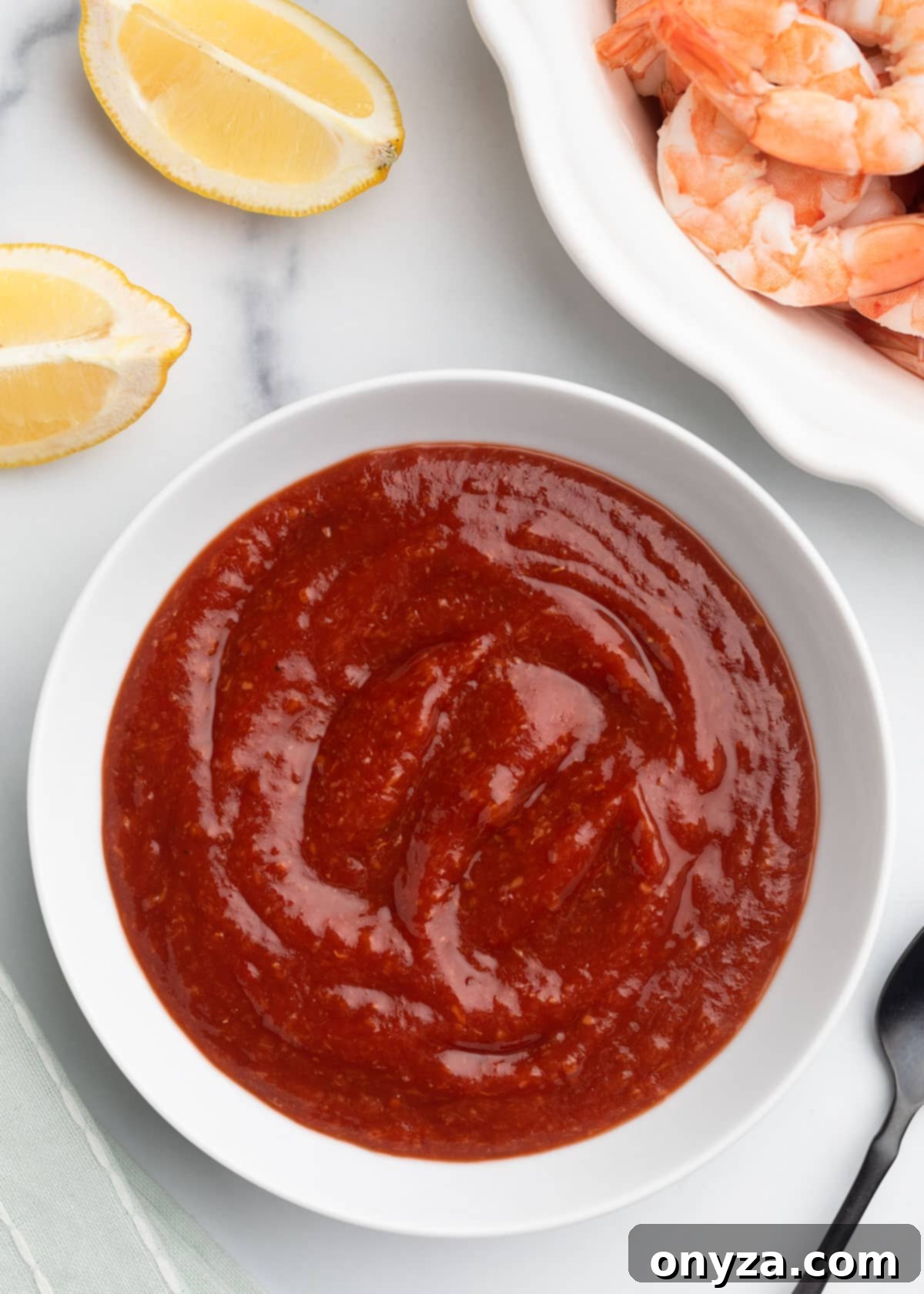 Zesty Homemade Cocktail Sauce 2 Overhead shot of vibrant homemade cocktail sauce in a pristine white bowl, elegantly arranged alongside bright lemon wedges, a platter of cooked shrimp, and a sleek black serving spoon.