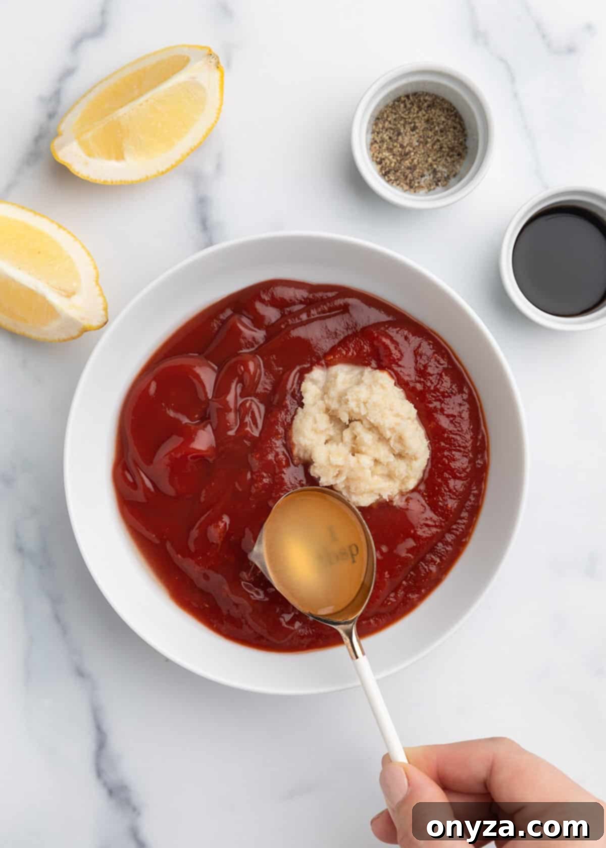 Zesty Homemade Cocktail Sauce 4 Overhead photo of fresh lemon juice being poured from a tablespoon into a white bowl containing unmixed homemade cocktail sauce, set on a elegant marble board.