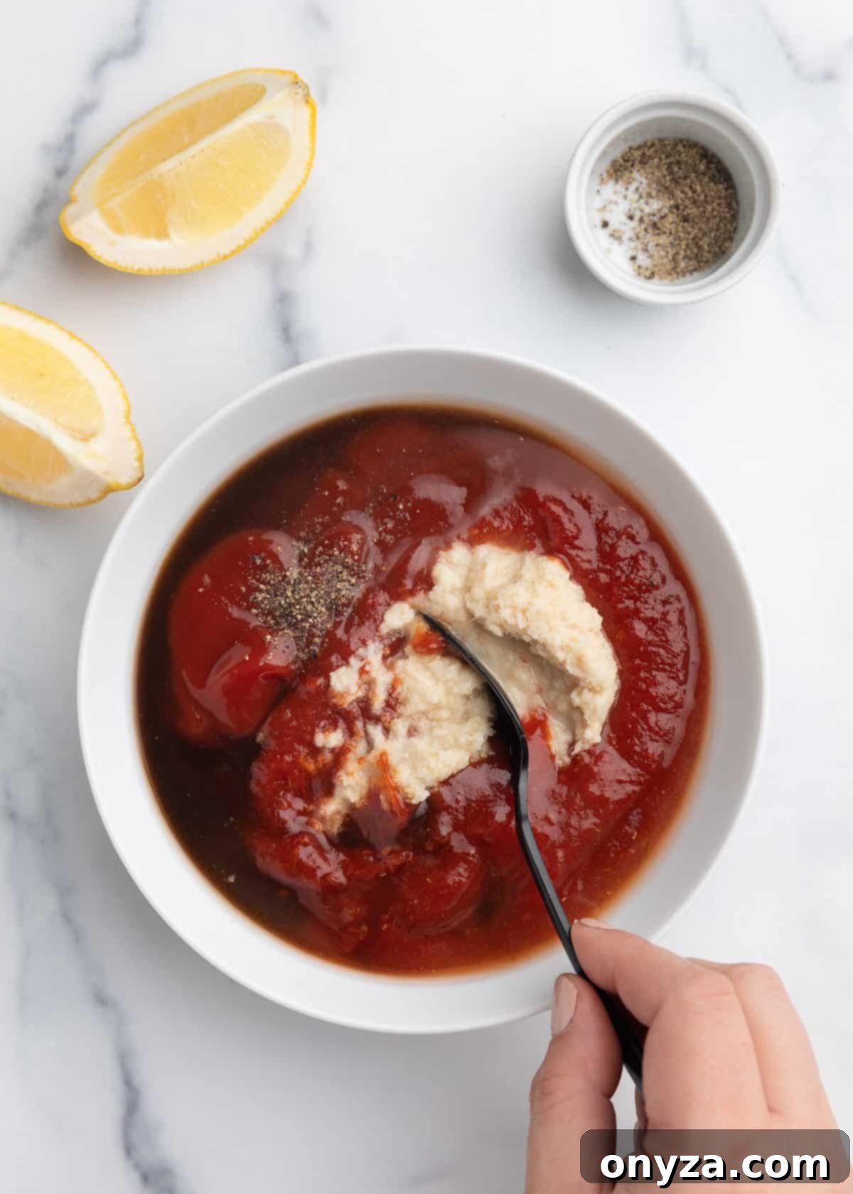 Zesty Homemade Cocktail Sauce 5 Overhead photo of the various ingredients for cocktail sauce being gently stirred together in a white bowl, resting on a sophisticated marble board.