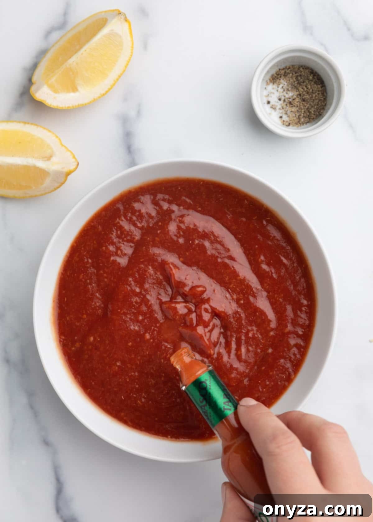 Zesty Homemade Cocktail Sauce 6 A bottle of hot sauce being shaken, adding a few dashes into a bowl of homemade cocktail sauce, all placed on a pristine white marble board.