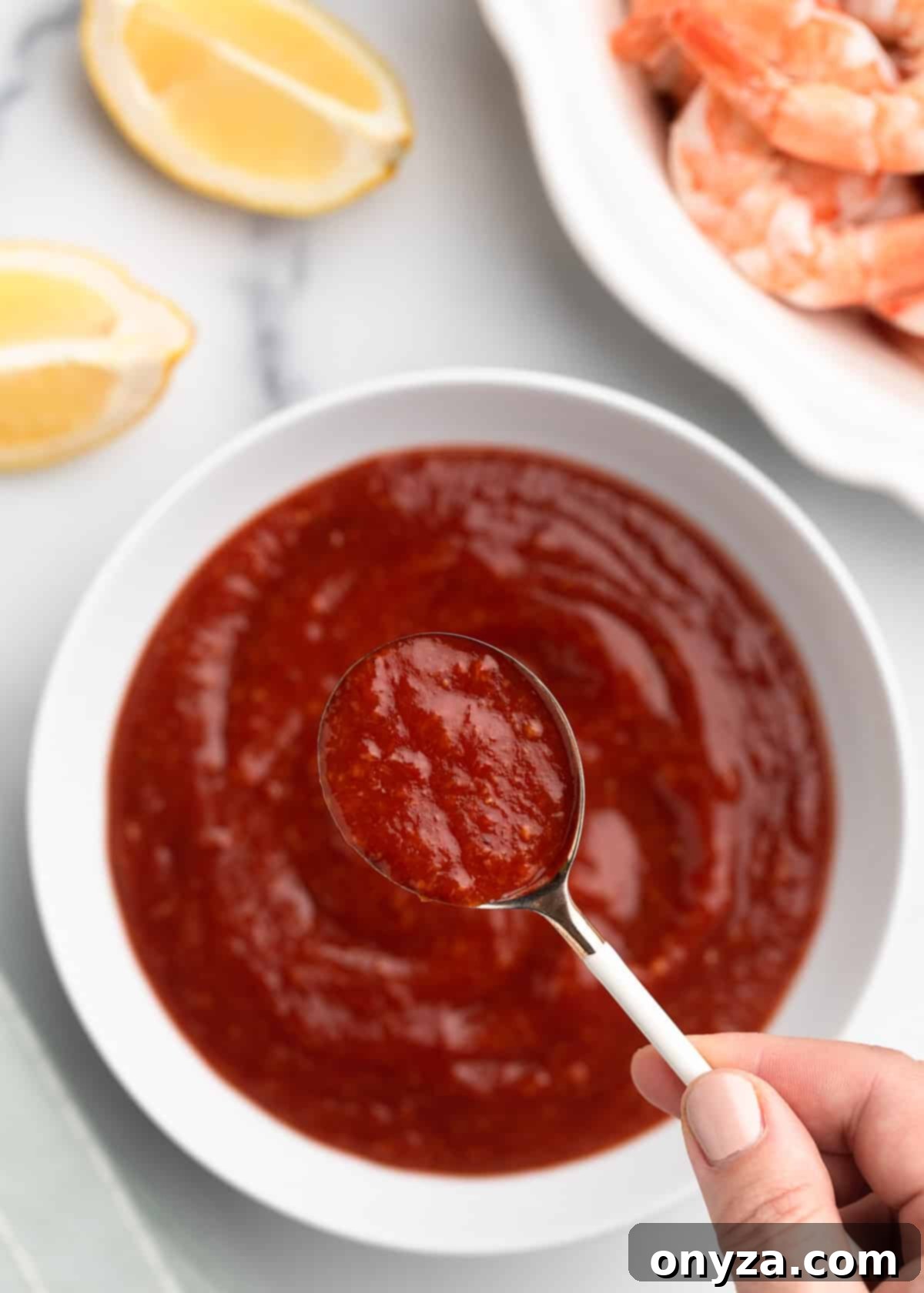 Zesty Homemade Cocktail Sauce 8 Overhead photo of a spoonful of homemade cocktail sauce being held tantalizingly over a white bowl of the sauce, placed on a white board beside a plate of plump shrimp and bright lemon wedges.
