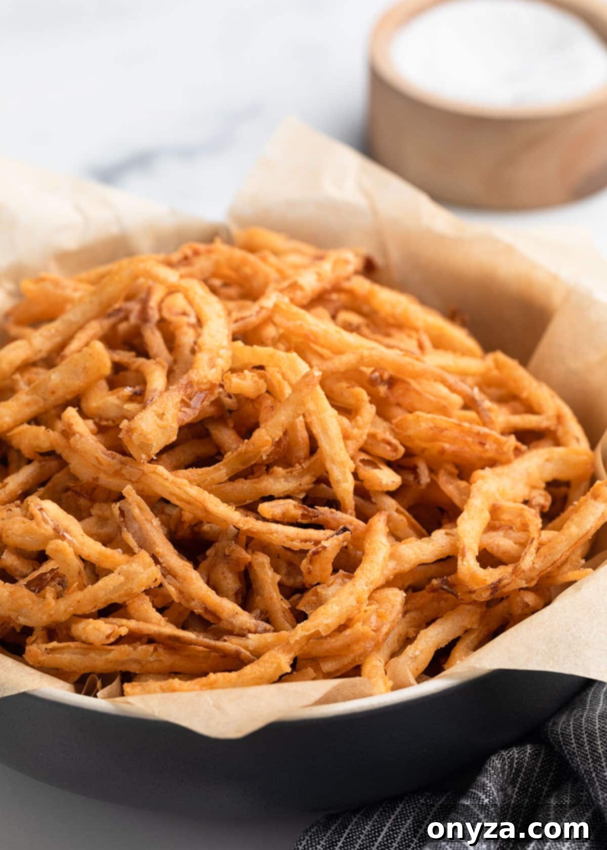 A close-up of a black bowl lined with parchment paper, generously filled with golden, crispy homemade French fried onions, perfectly showcasing their inviting texture. In the softly blurred background, a gray striped napkin and a wooden salt cellar hint at a cozy kitchen setting.