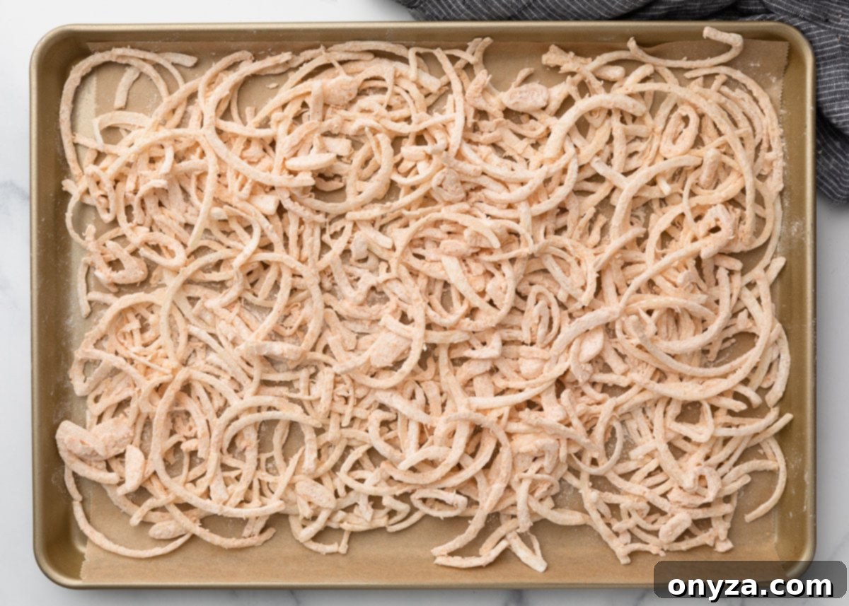 A baking sheet generously lined with parchment paper, displaying a single, neat layer of thinly sliced onions, each piece perfectly coated in the seasoned flour mixture. They are meticulously arranged and ready for the deep frying stage to achieve golden perfection.