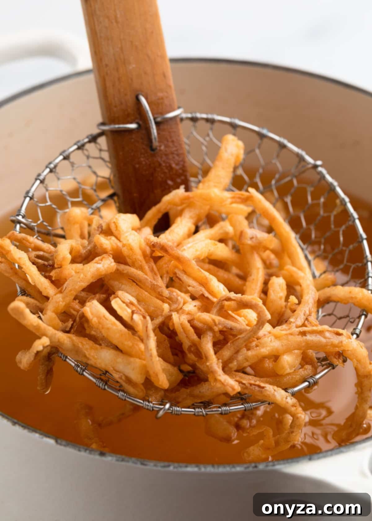 A cluster of perfectly golden brown French fried onions resting gracefully in a wire spider skimmer, held just above a pot of hot oil, having just been expertly lifted from frying. This image proudly showcases their perfect crispness and appealing golden color.