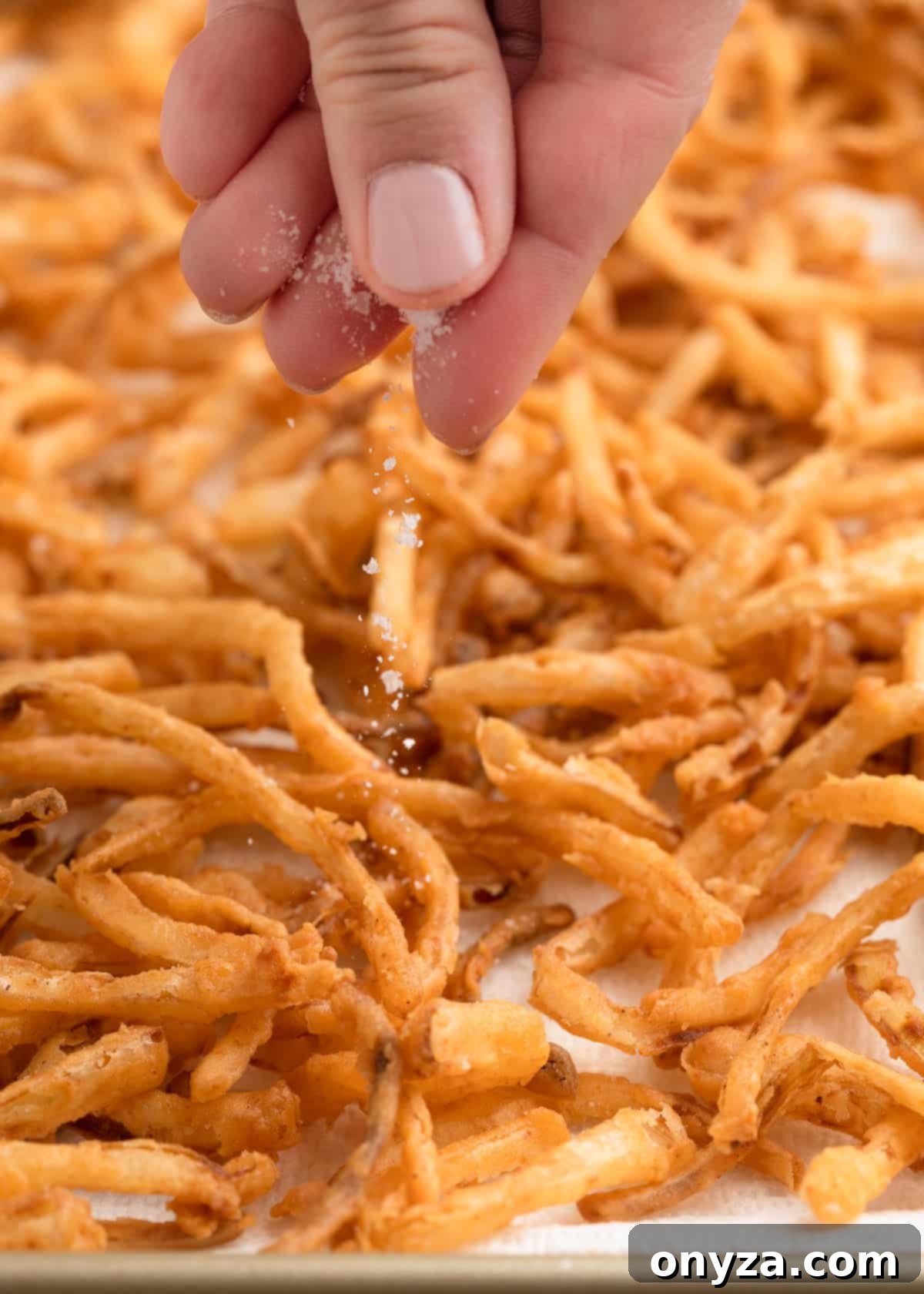 A close-up shot of a hand meticulously sprinkling coarse salt over a tray of freshly fried, crispy homemade onions. The onions are spread out on parchment paper to drain, vividly capturing the final, crucial seasoning step.