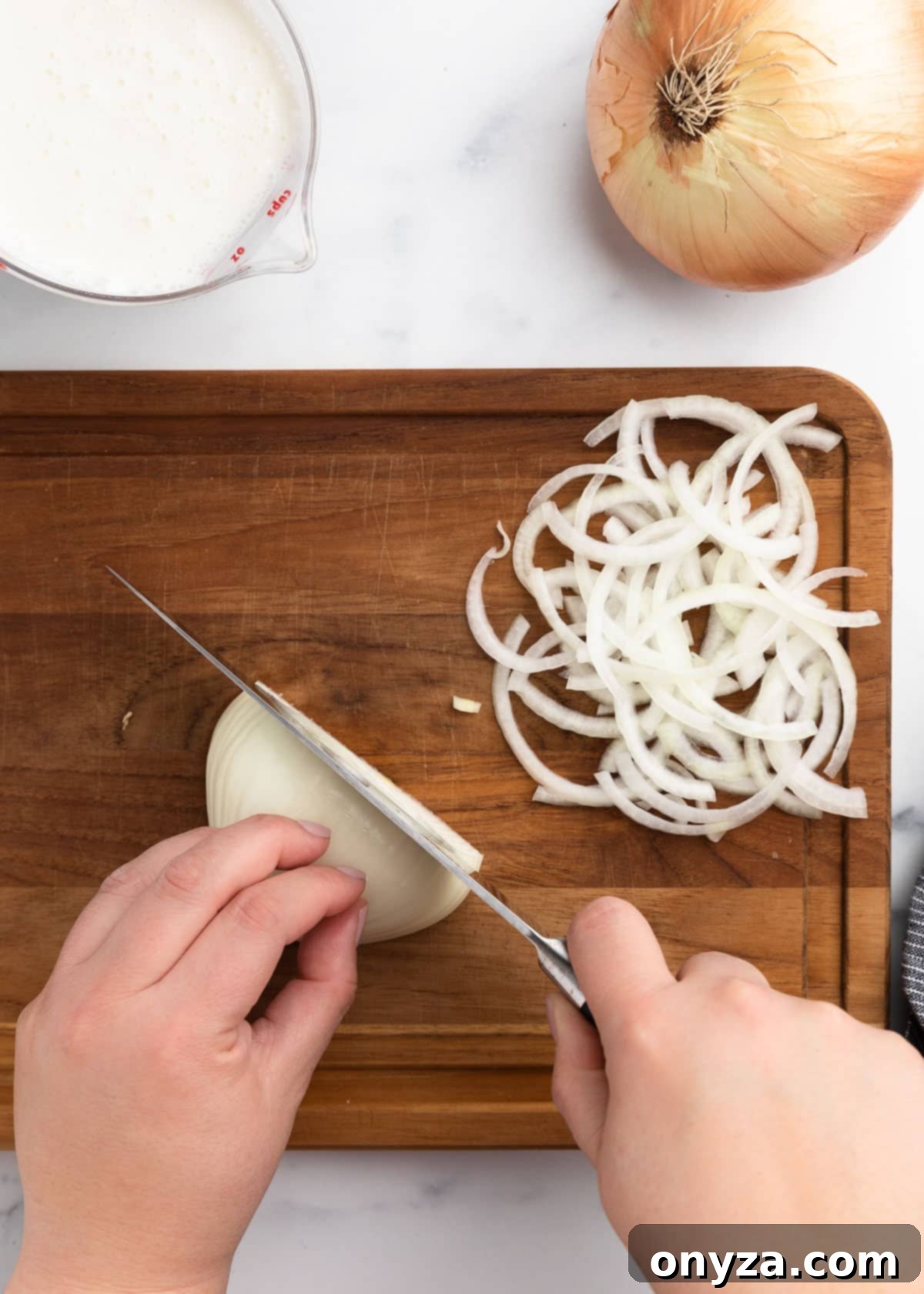 A pair of hands carefully slicing a sweet onion into thin half-moons on a wooden cutting board, with a small pile of already thinly sliced onion rings nearby, demonstrating the meticulous cutting technique required.