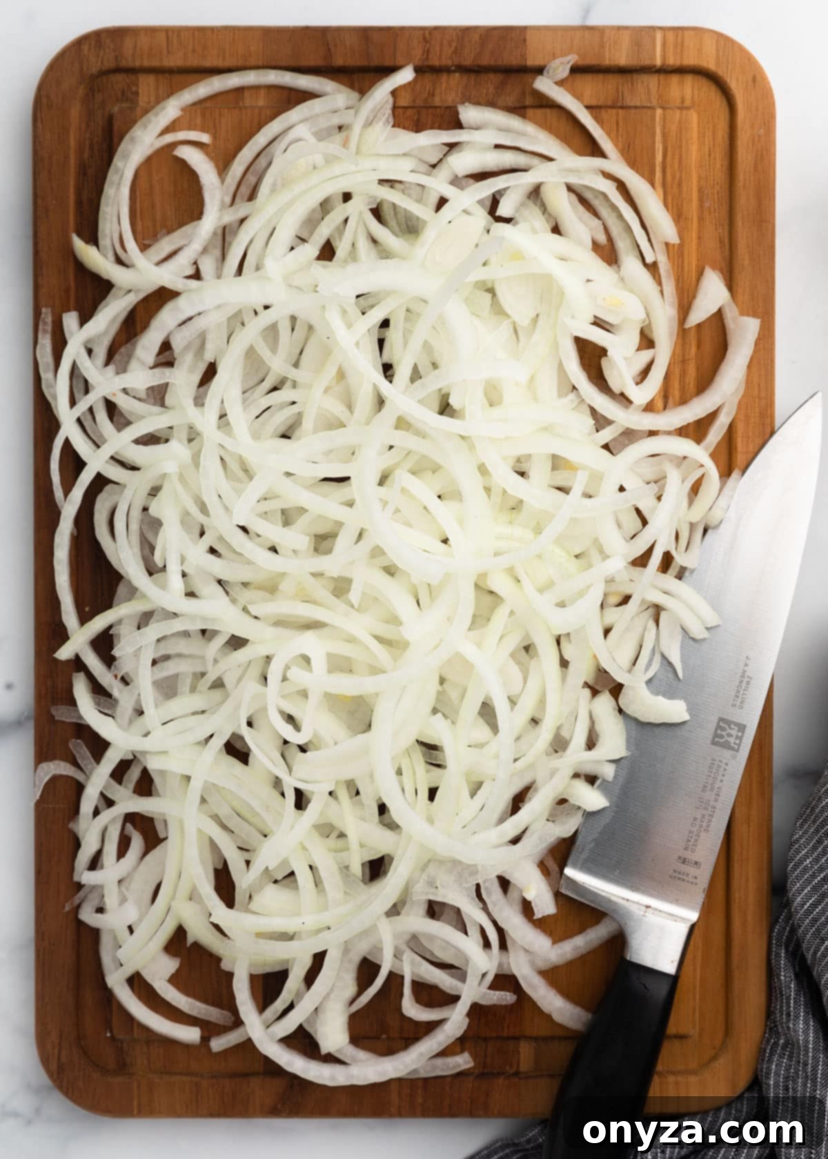 A cutting board generously piled with thinly sliced white onion rings, neatly arranged and separated, with a chef’s knife resting alongside and a striped kitchen towel in the corner, signaling readiness for the crucial soaking step.