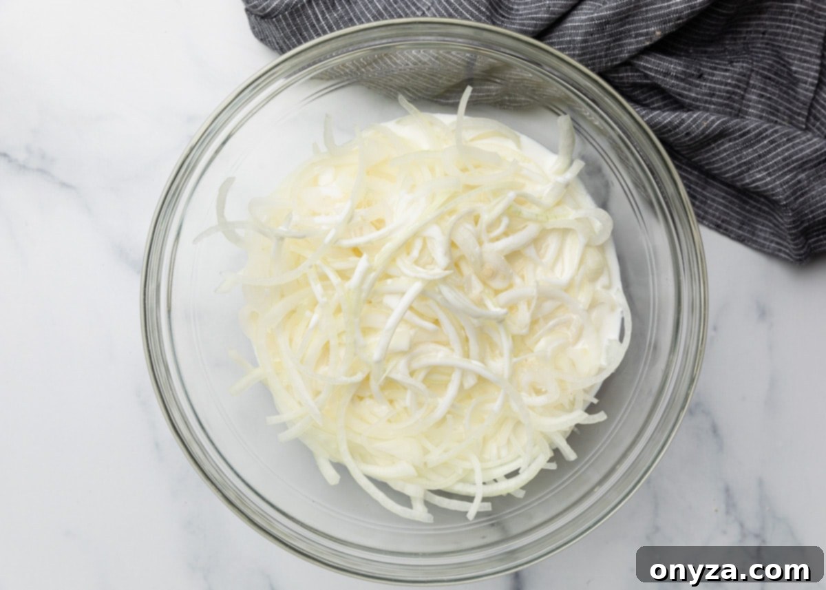 Thinly sliced onions luxuriating in a clear glass bowl, fully submerged in creamy buttermilk. The bowl rests on a pristine marble surface, with a chic gray striped towel nearby, visually emphasizing this vital tenderizing and coating preparation step.