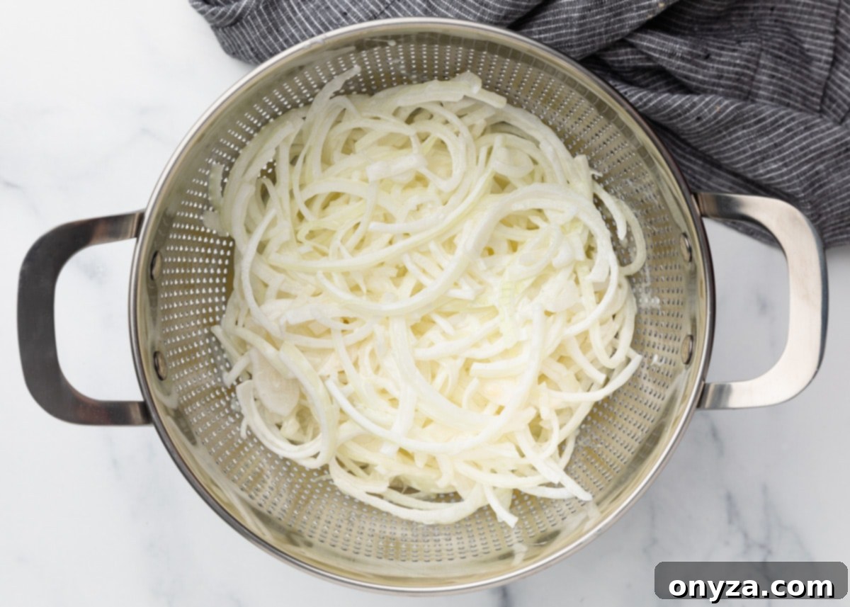 A cluster of sliced onions diligently draining in a sleek metal colander, positioned over a pristine marble surface. A crisp gray striped kitchen towel is neatly folded to the side, underscoring the vital importance of thorough drainage before the next step.