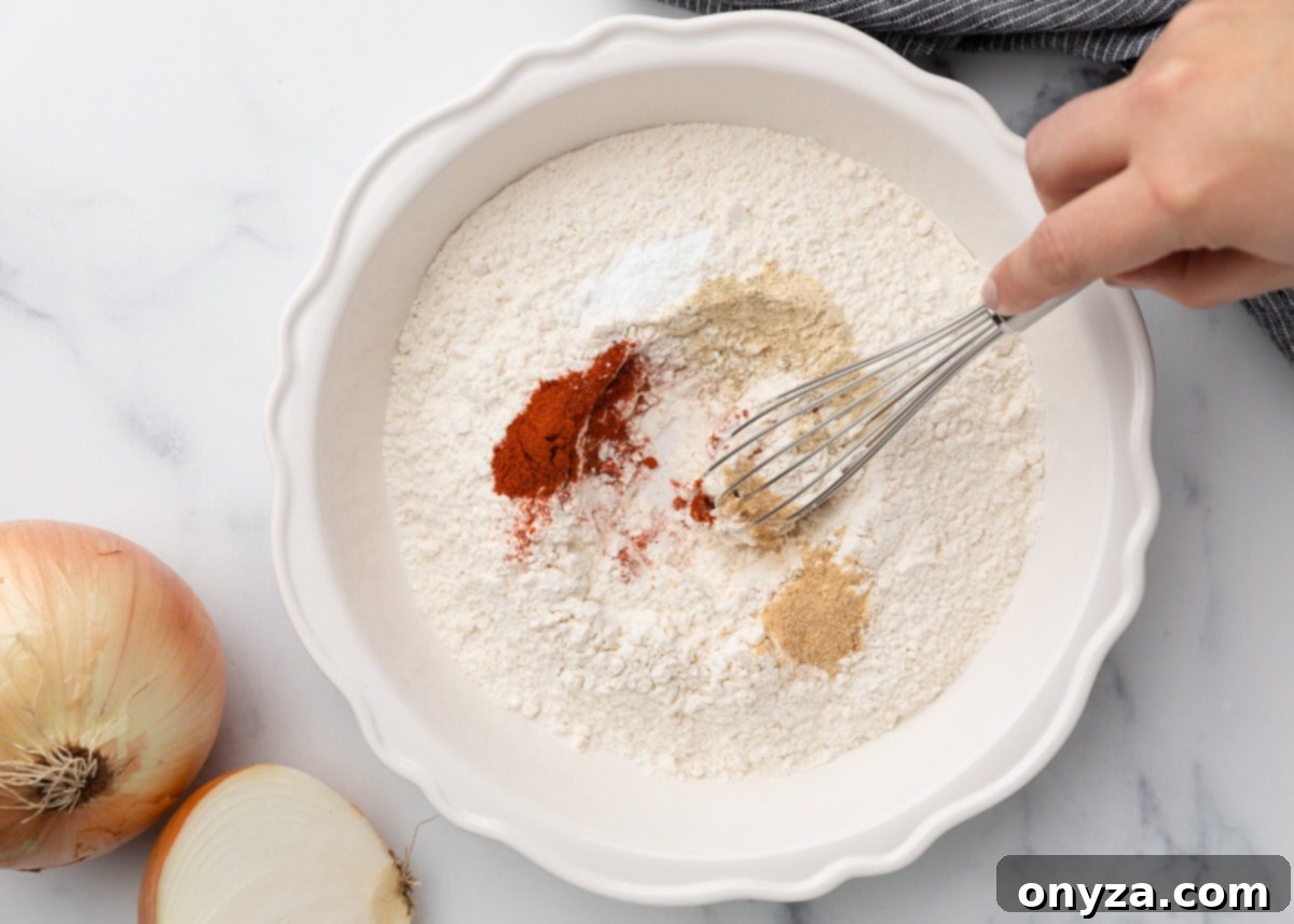 A pristine white ceramic bowl, showcasing an ideal blend of all-purpose flour, baking powder, and a vibrant array of spices, including reddish paprika and fine garlic powder. A hand is actively whisking this seasoning blend together, preparing the perfect coating for the onions.