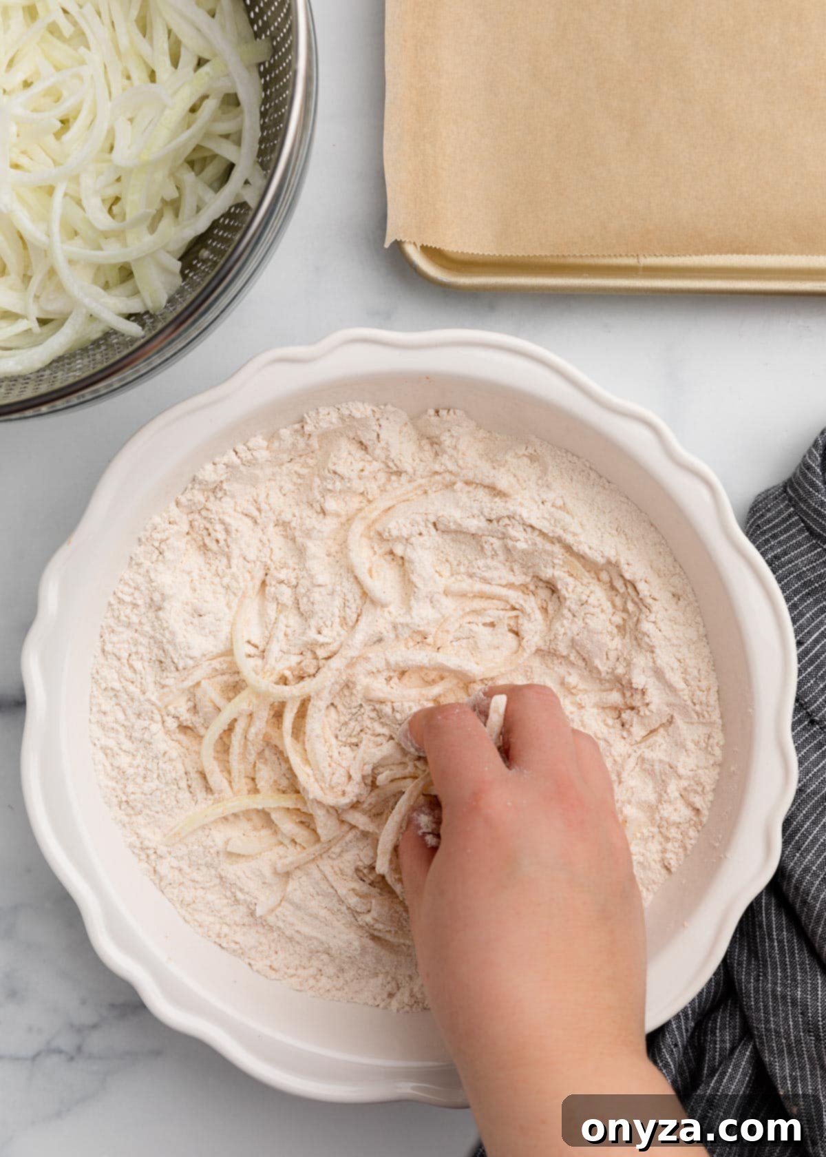 A hand gently tossing thinly sliced onions in the seasoned flour mixture within a white bowl. In the background, a metal colander holds remaining drained onions, and a parchment-lined baking sheet awaits the coated pieces, perfectly illustrating the careful coating process.