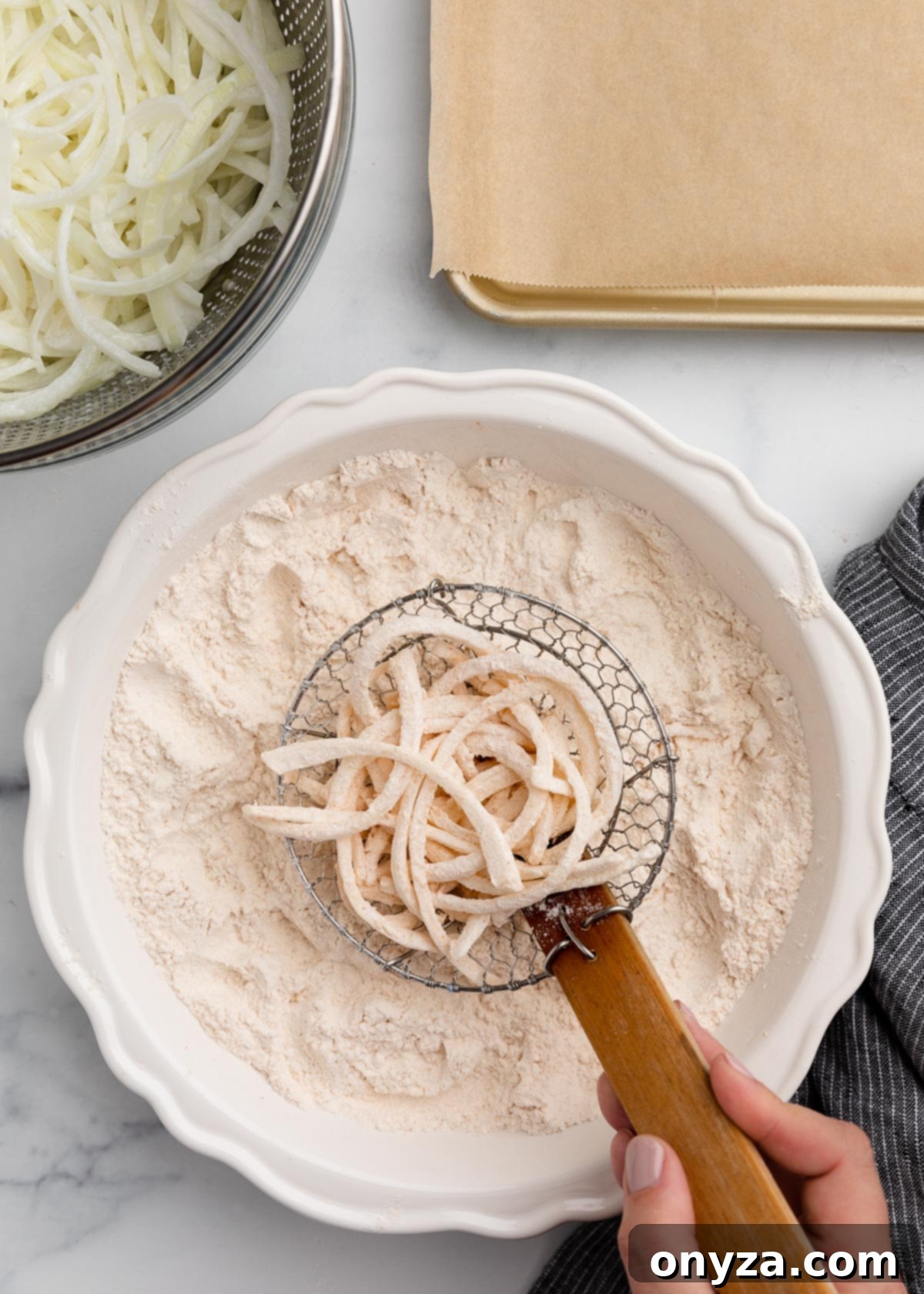 A wire spider strainer deftly holding a portion of flour-dredged onion slices directly over a bowl of seasoned flour, with a metal colander of fresh onions and a prepared baking sheet in the periphery, clearly demonstrating the technique to remove excess flour.