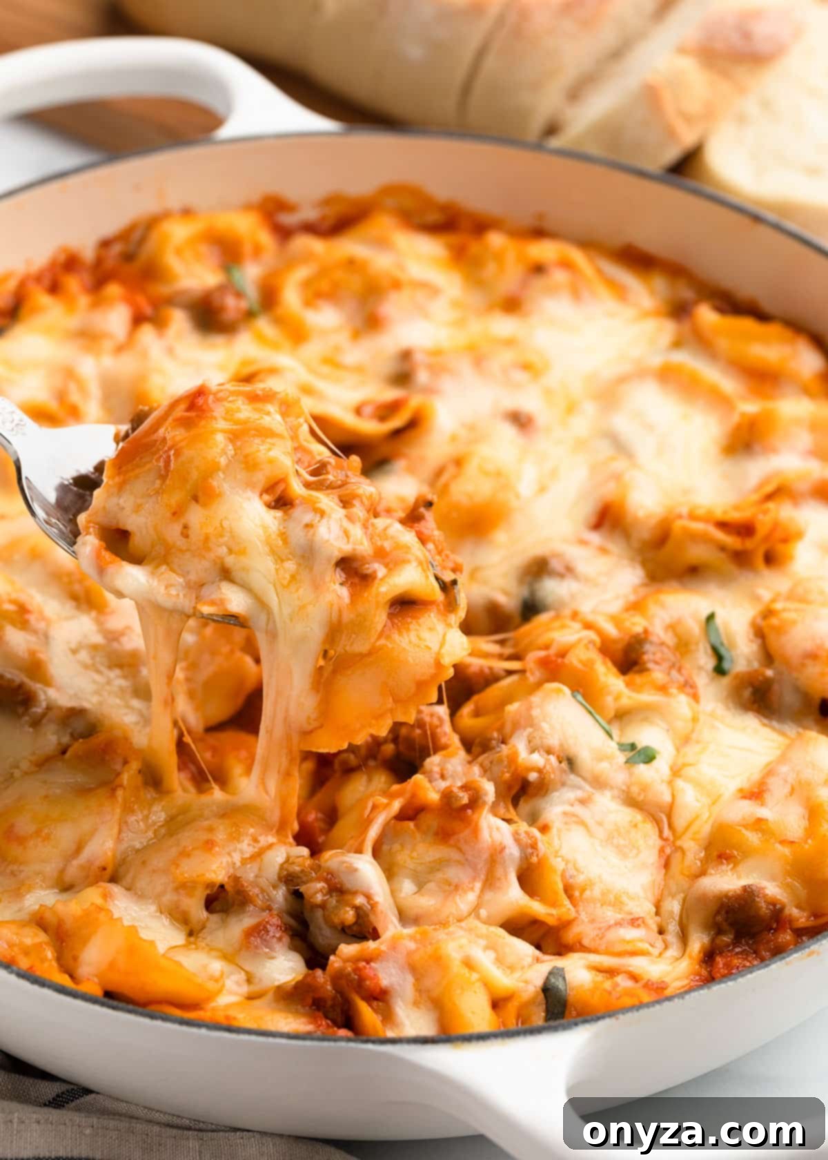 A spoonful of baked tortellini with sausage being scooped from a white enameled cast iron pan with a satisfying cheese pull.