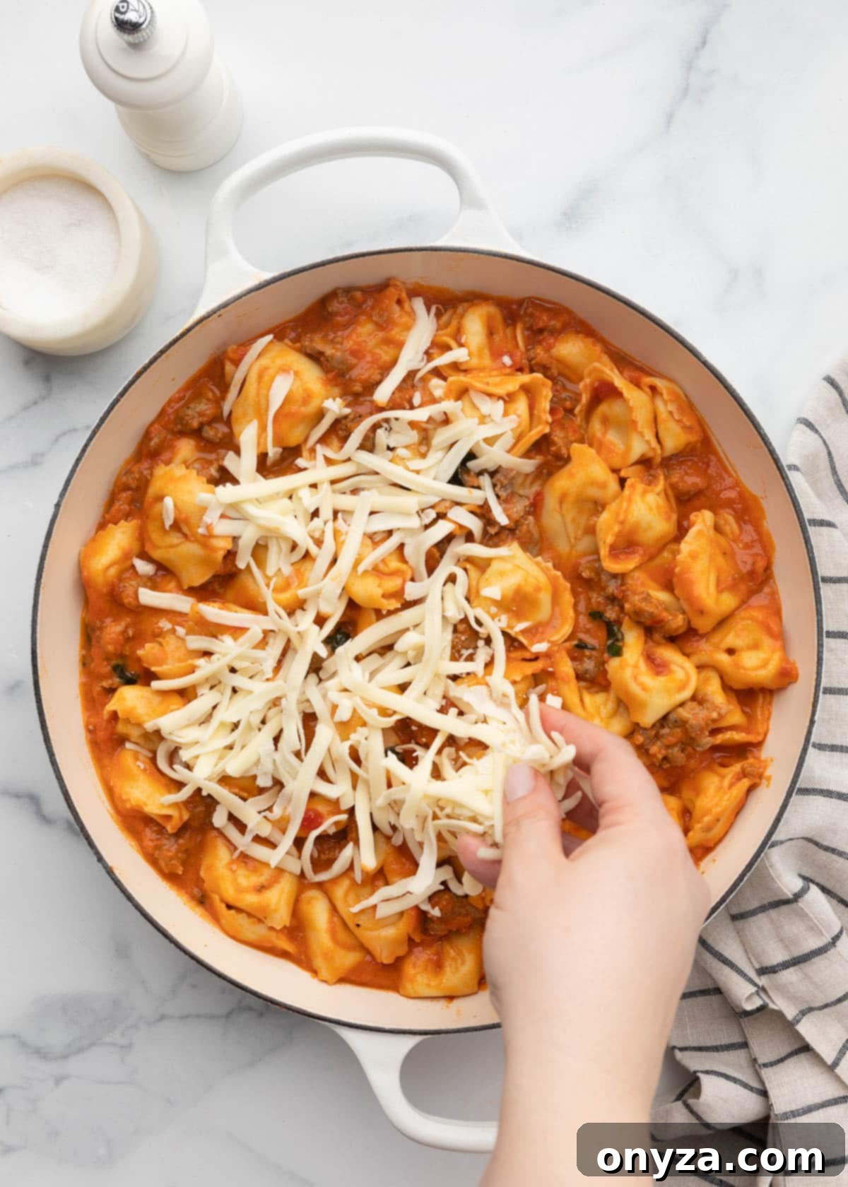 Sprinkling shredded mozzarella generously over the sauced tortellini in a white enameled cast iron pan.
